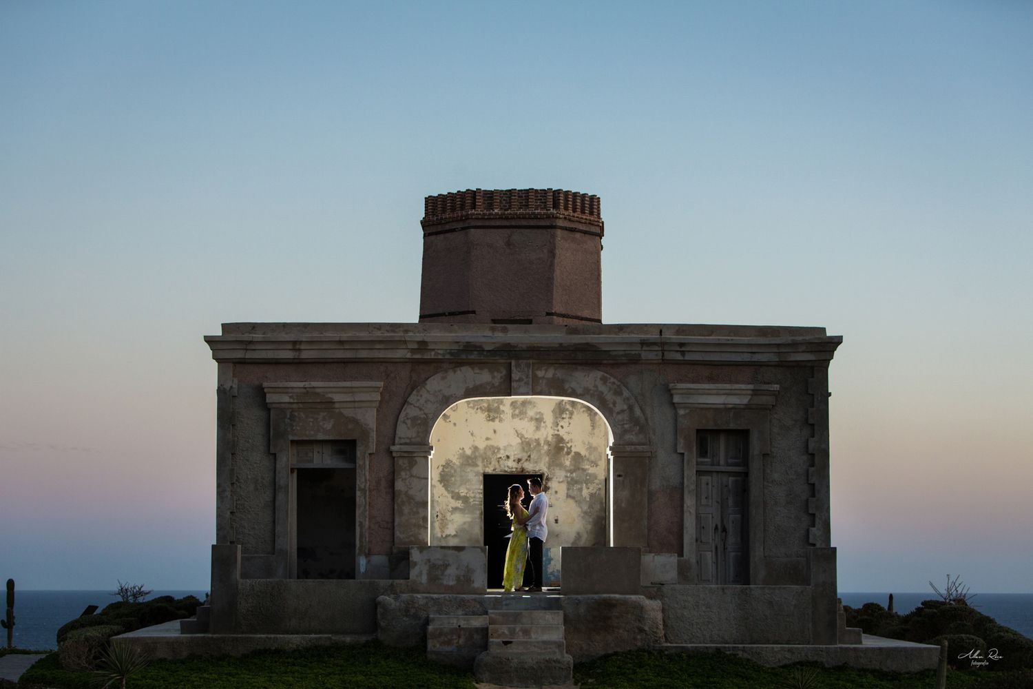 Couple embracing inside an arched stone monument at dusk with ocean in background.