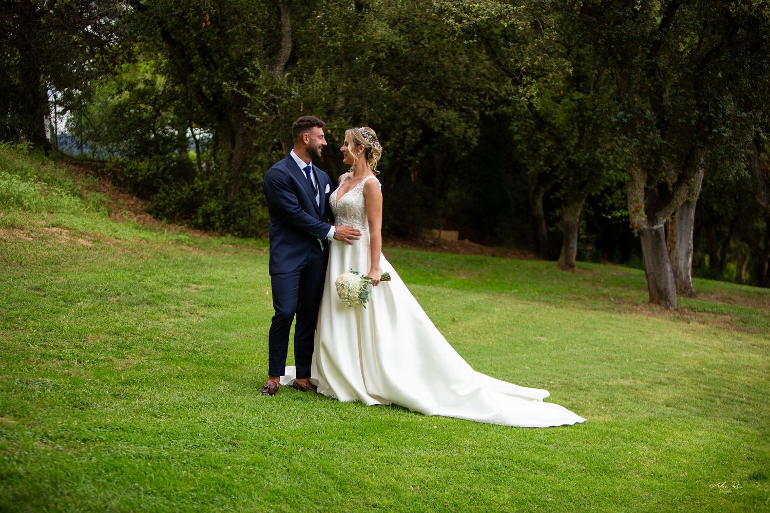 Bride and groom share a romantic moment on lush green grass surrounded by trees outdoors.