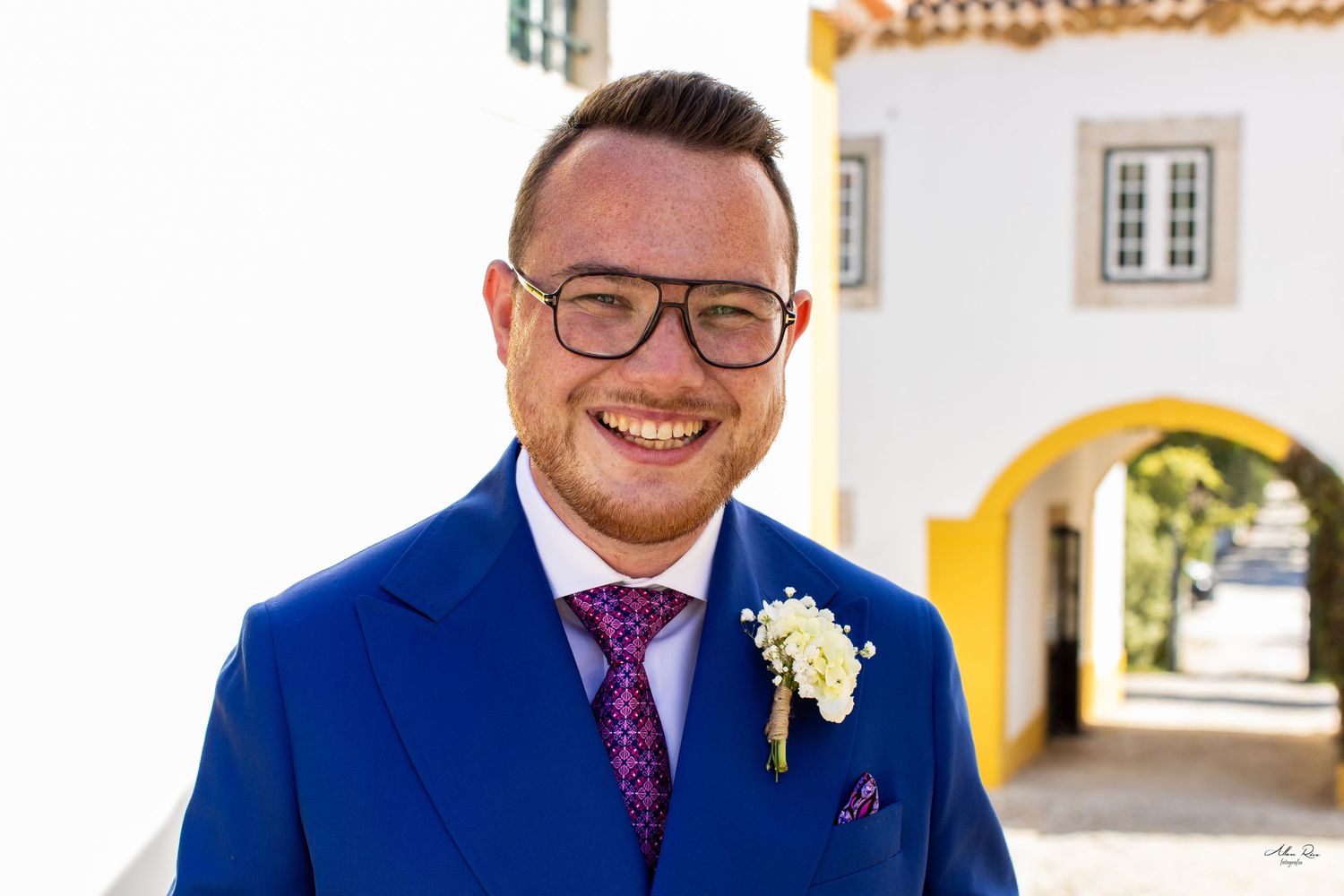 Smiling groom in blue suit with floral boutonniere at sunny outdoor venue.