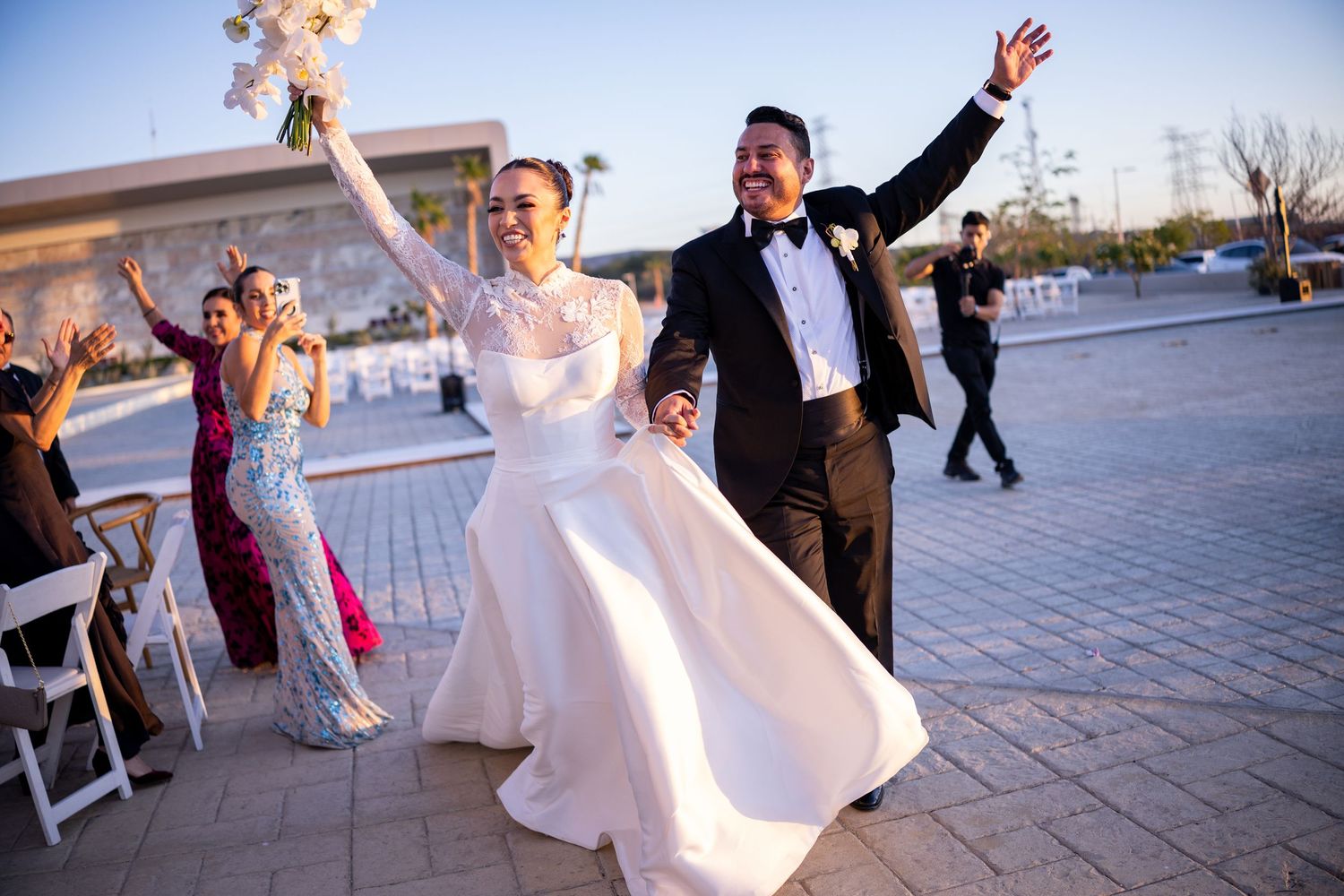 Joyful newlyweds celebrate wedding exit with raised hands and bouquet on outdoor venue patio.