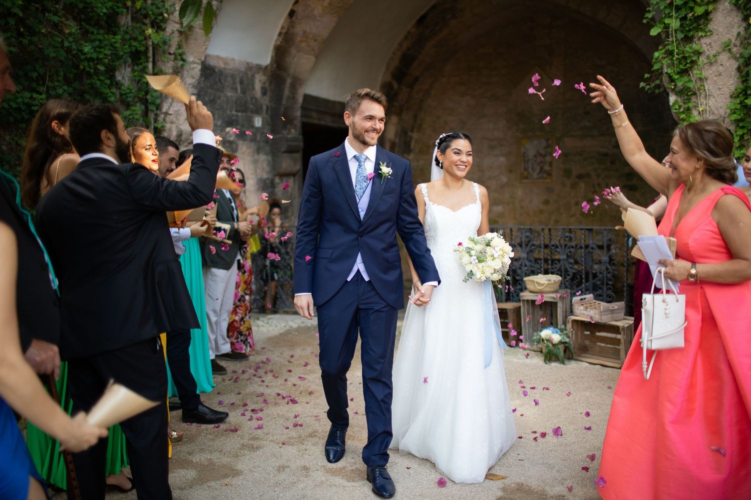 Bride and groom walking through flower petal confetti shower during joyful outdoor wedding ceremony exit.