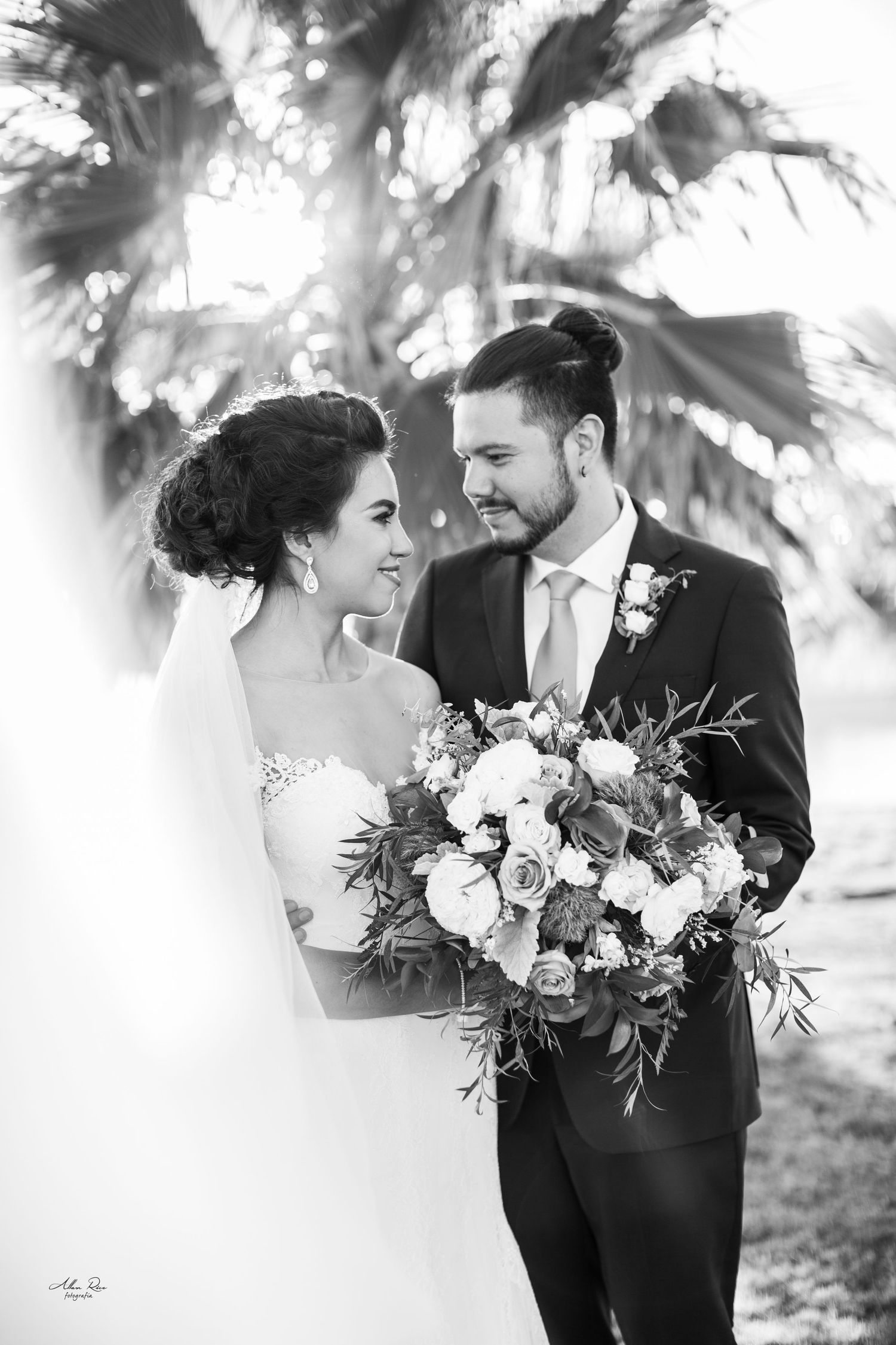 Bride and groom share a romantic moment under palm trees, bride holding a lush white and green bouquet.