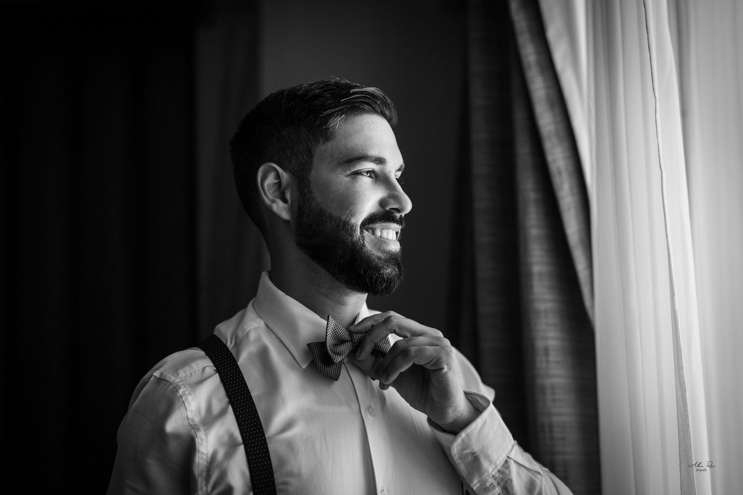 Smiling groom adjusting bow tie wearing suspenders in black and white wedding portrait by window.
