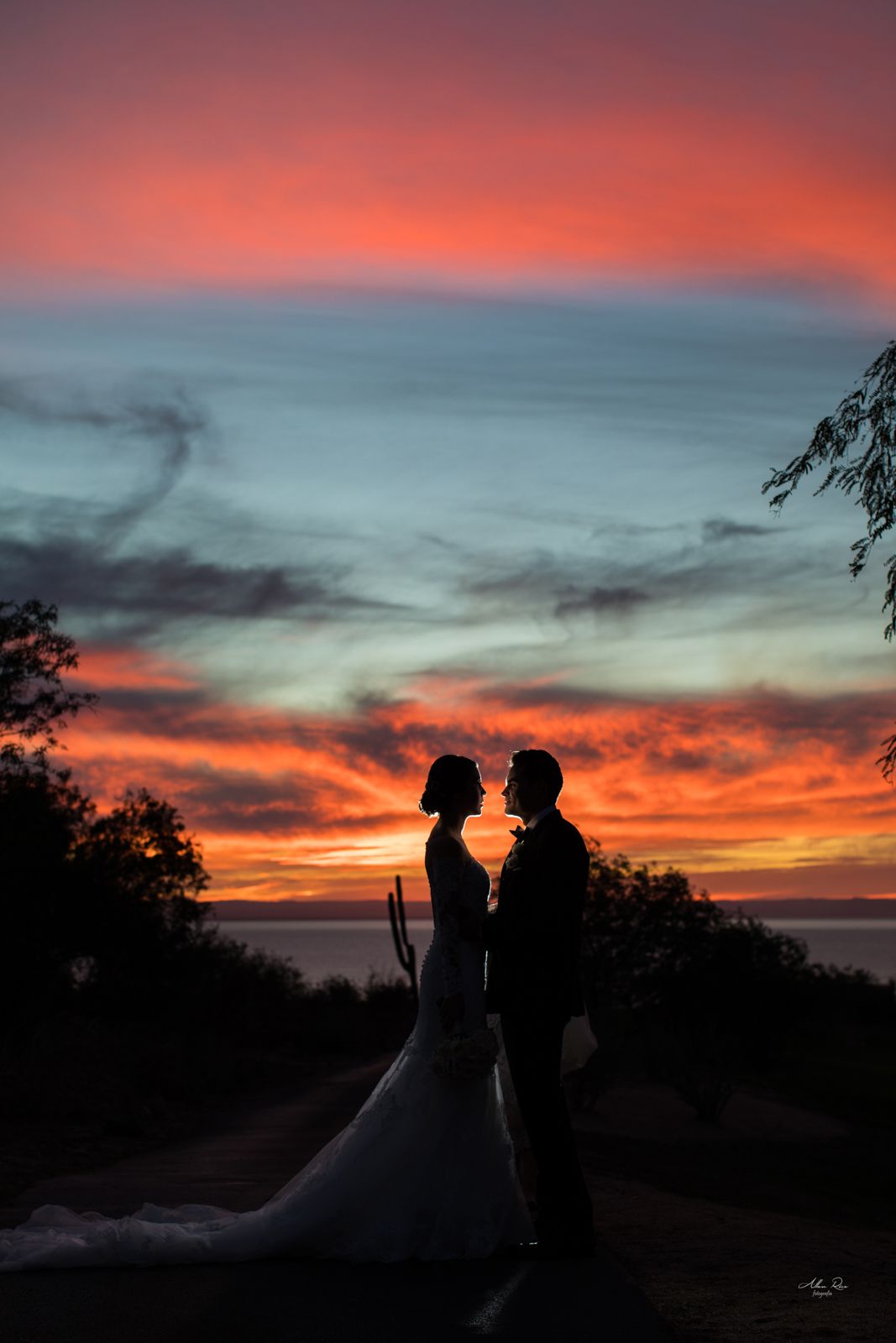 Silhouette of bride and groom facing each other against dramatic colorful sunset sky at outdoor wedding.