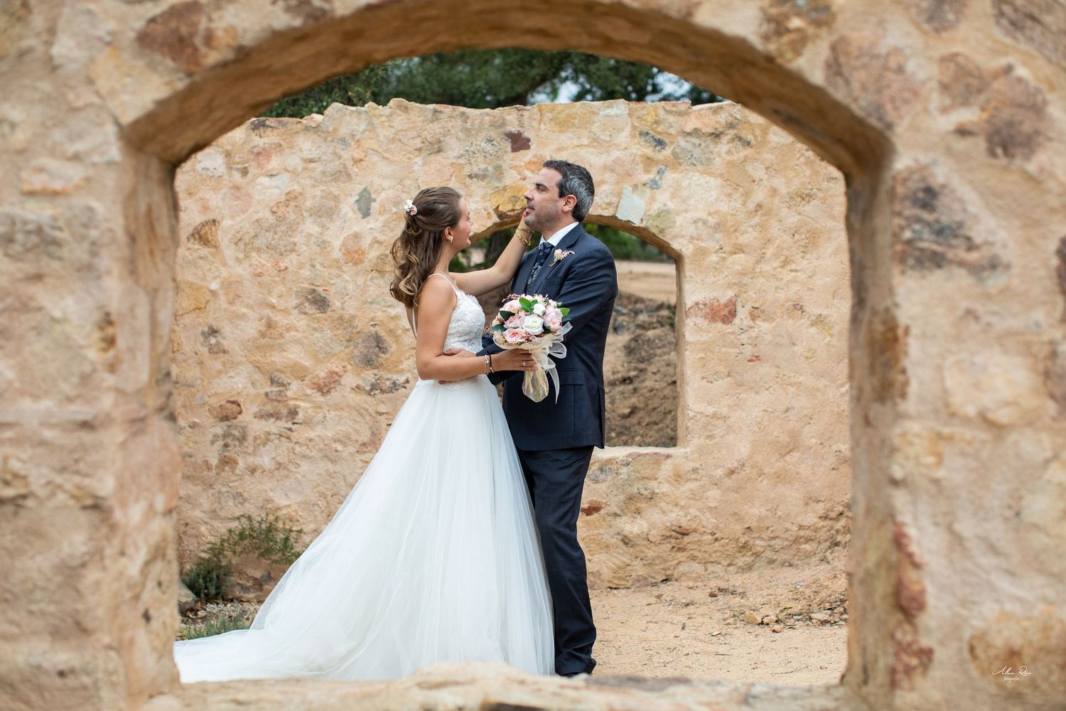 Bride and groom share tender moment framed by rustic stone archway outdoors.