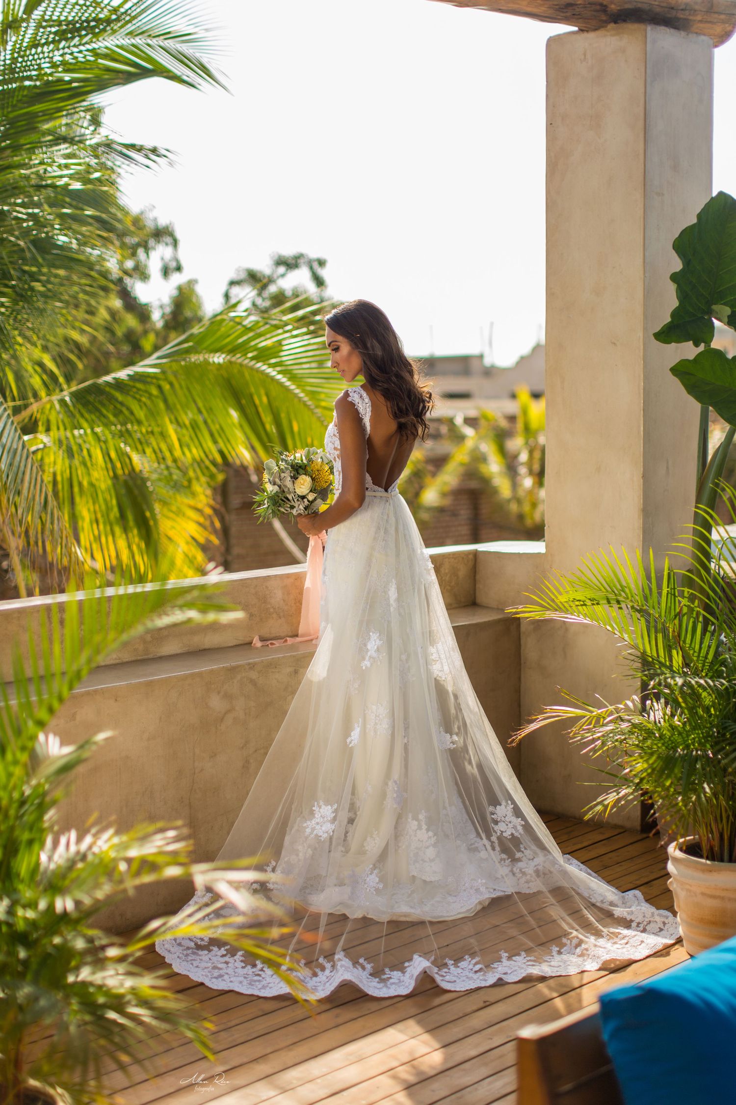 Bride in flowing lace gown with train holds bouquet on sunlit tropical terrace with palm trees.