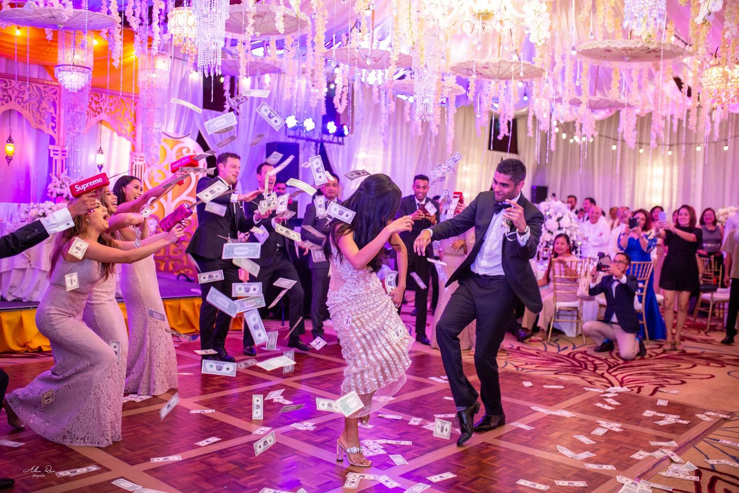 Guests celebrate on a dance floor throwing dollar bills at an elegantly decorated wedding reception with purple lighting.