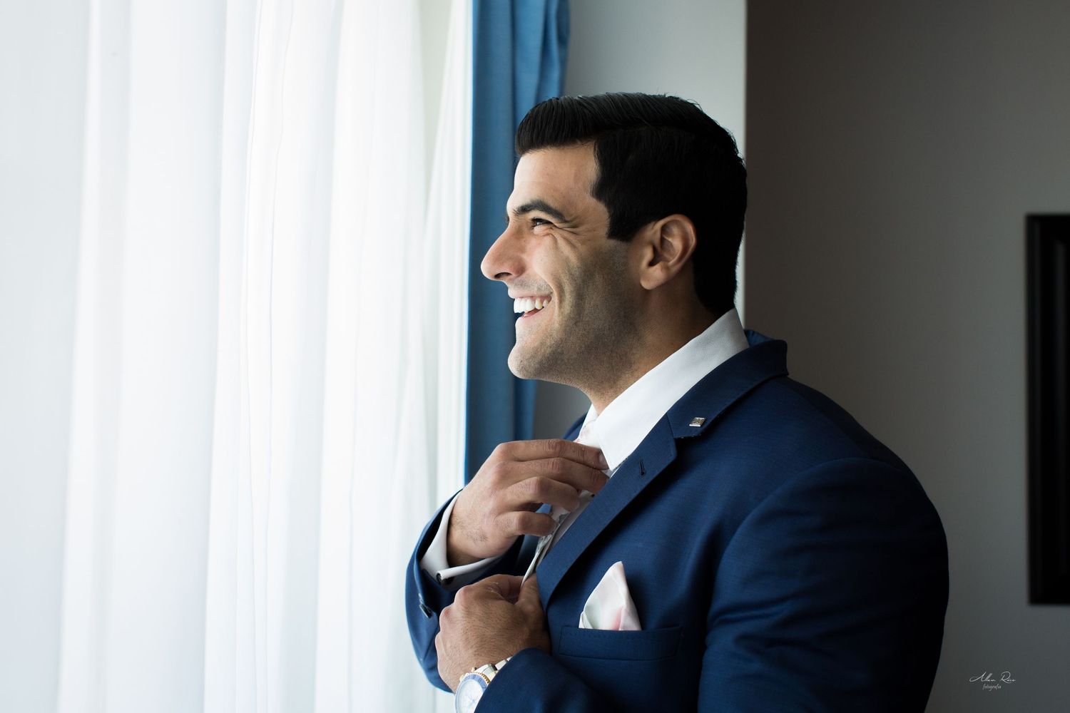 Handsome groom in navy blue suit smiling while adjusting his tie near a bright window on wedding day.