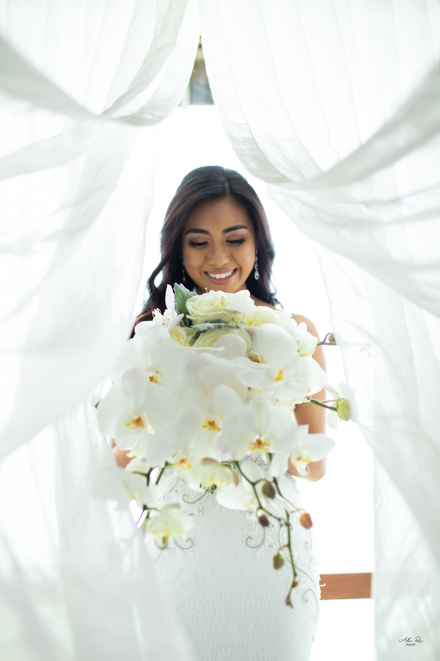 Joyful bride in white lace dress holding large cascading white orchid bouquet.