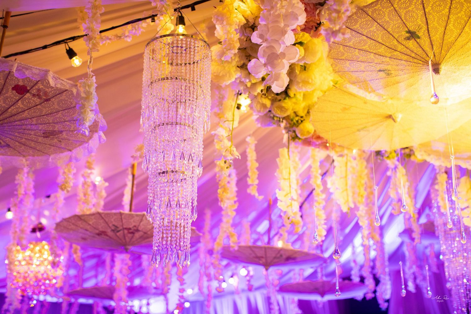 Lavish wedding reception decor with crystal chandeliers, white parasols, and purple ambient lighting.