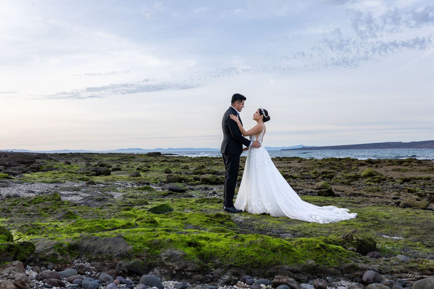 Bride and groom stand facing each other on moss-covered rocky beach with ocean horizon at dusk.