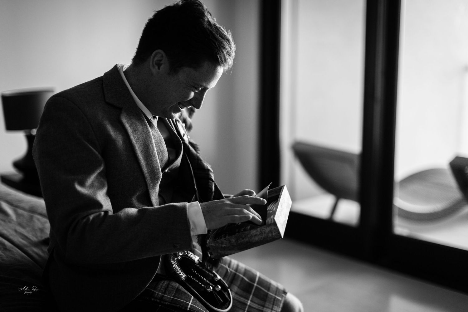 Groom in grey suit reads a heartfelt note while seated in a softly lit hotel room.