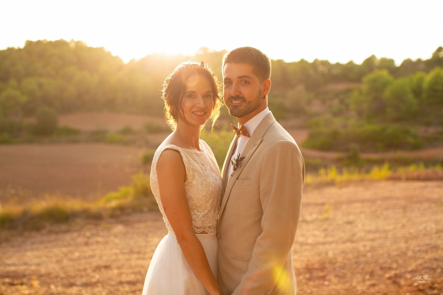 Smiling bride and groom posing together in a warm sunset field with lush green trees.