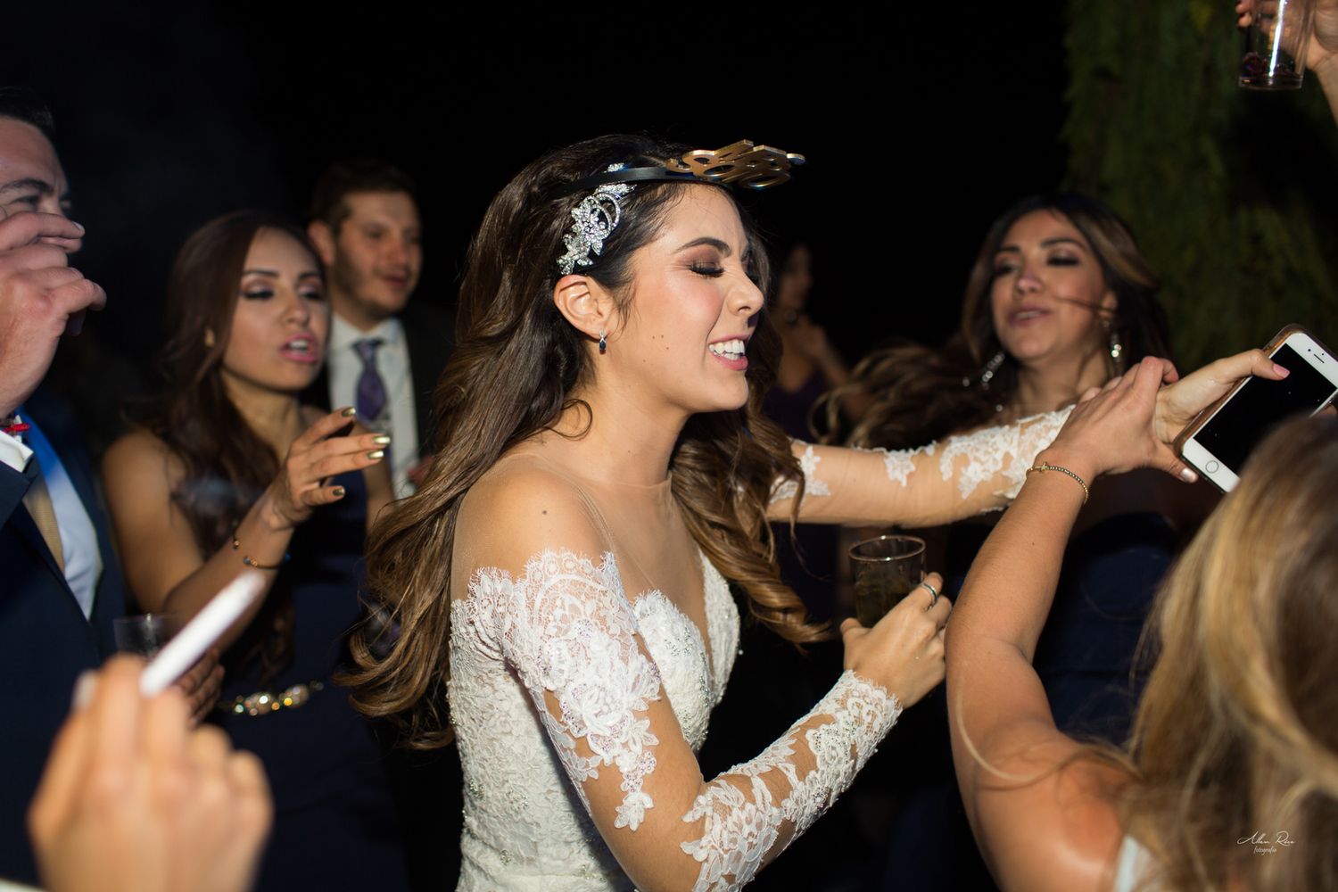 Bride in lace off-shoulder gown dancing joyfully with guests at evening wedding reception.