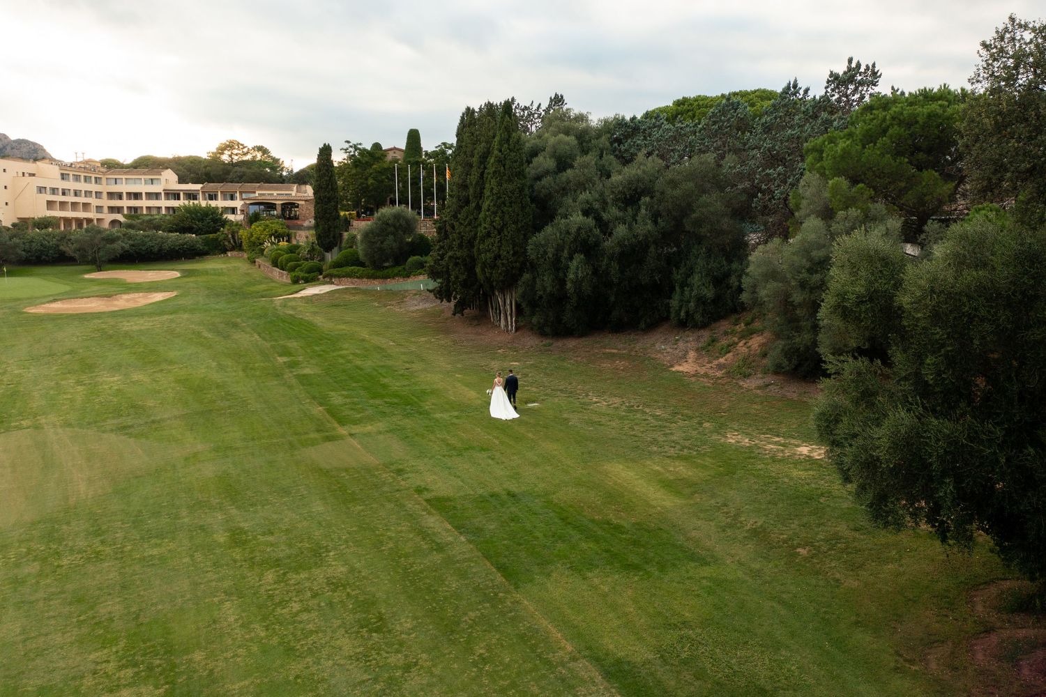 Aerial view of bride and groom standing on a golf course with a hotel building in background.