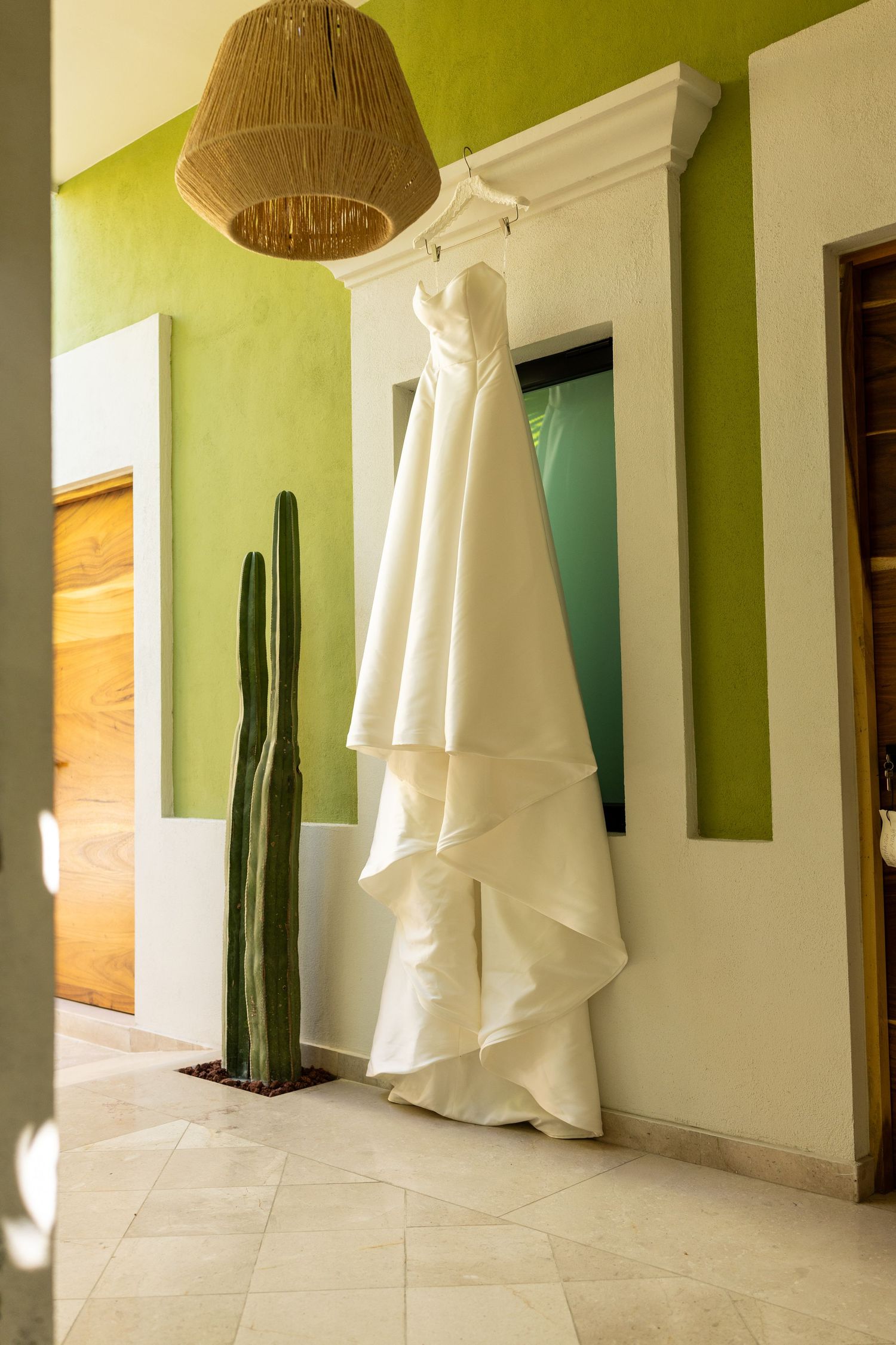 Strapless layered white wedding dress hanging on a door in a green-walled room with a cactus.