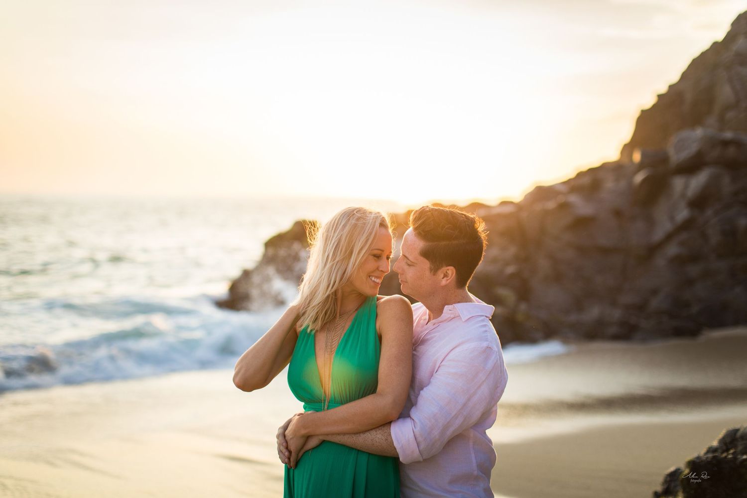 Couple sharing a romantic moment on a sandy beach at golden sunset with ocean waves and rocky cliffs.