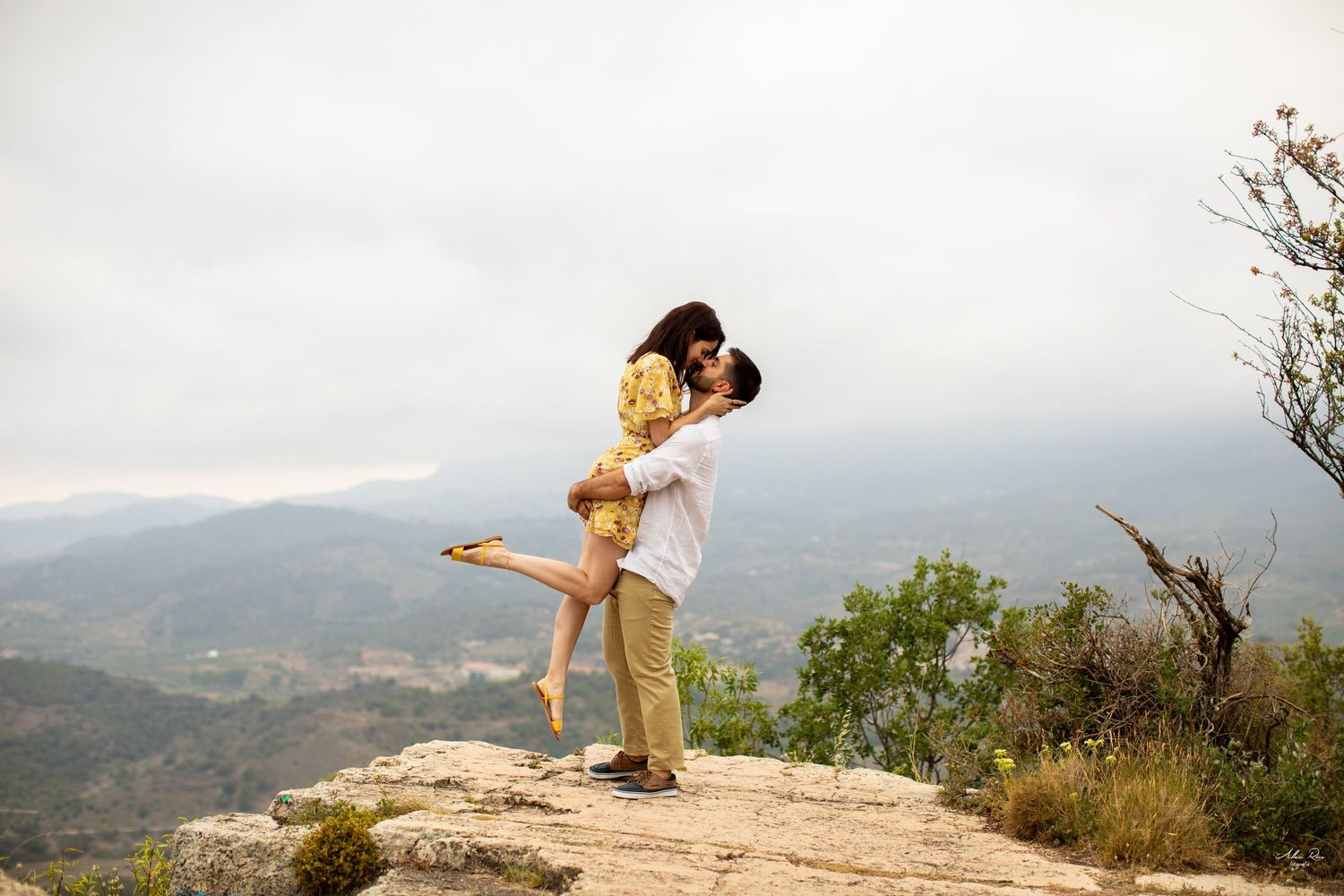 Man lifting and kissing woman on a rocky mountain summit with dramatic cloudy sky and valley views.