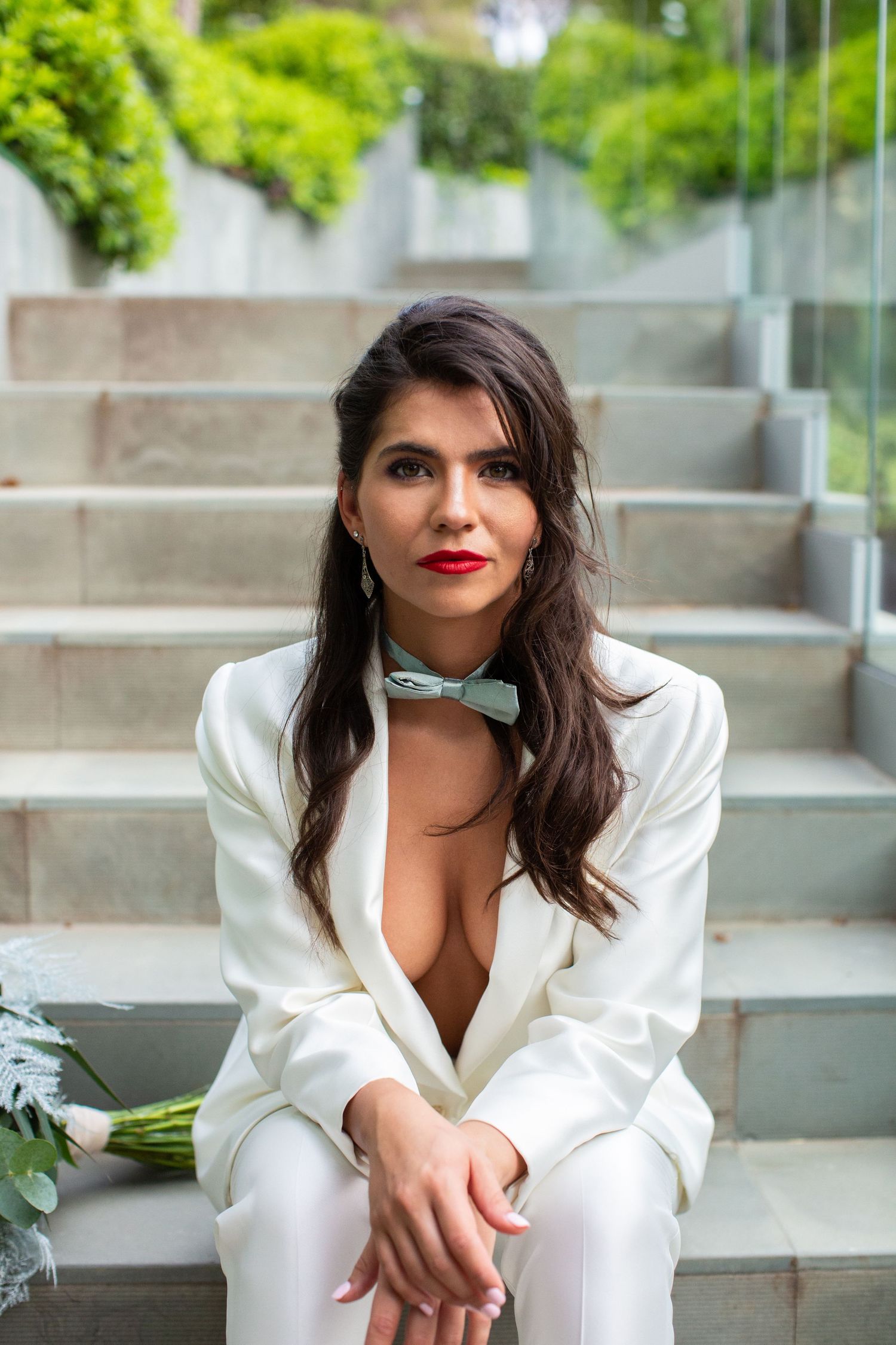 Stylish bride in a white suit with bow tie sits on stone steps holding a bouquet in a modern outdoor setting.