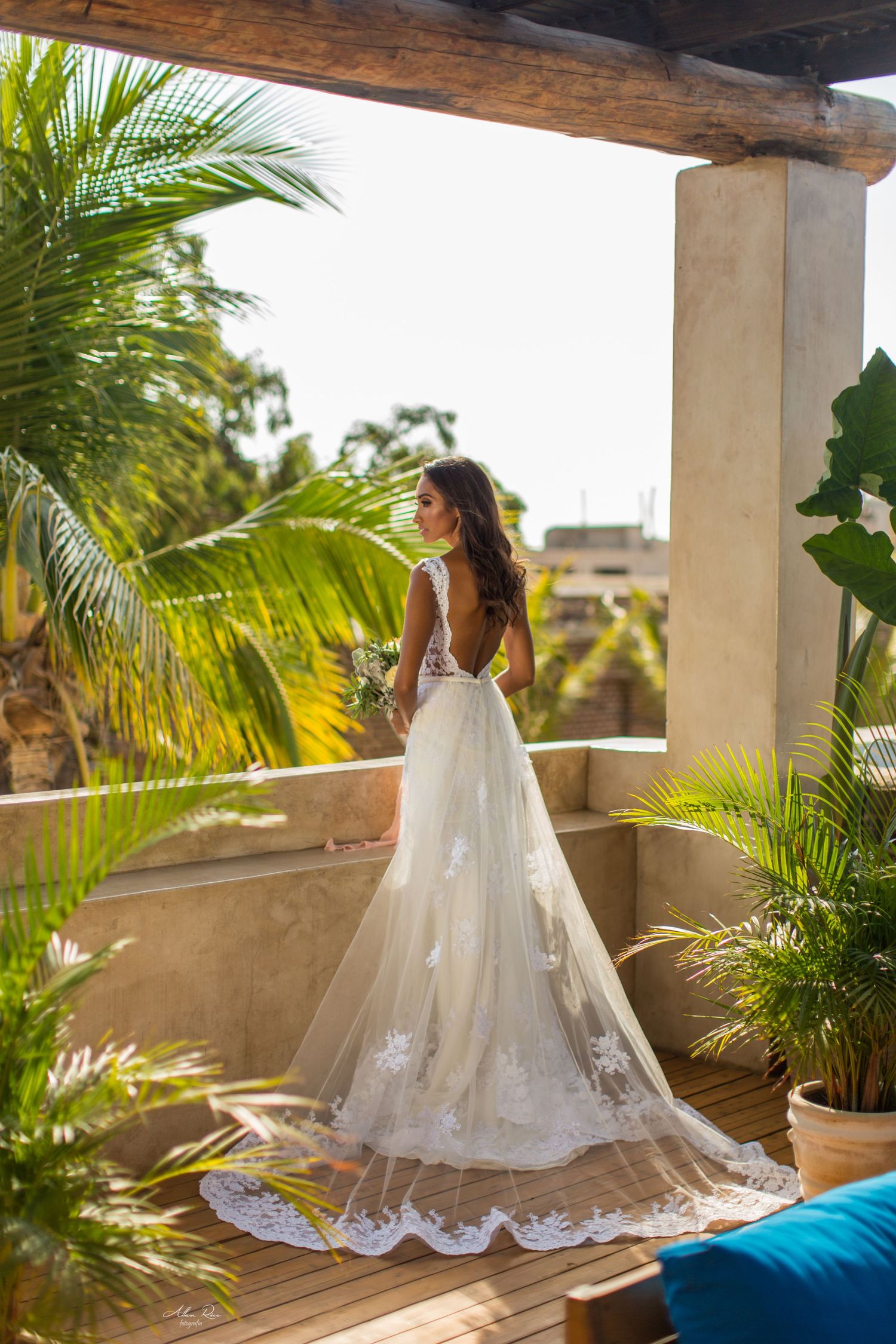 Elegant bride in backless lace wedding gown with long train on tropical terrace with palm trees.
