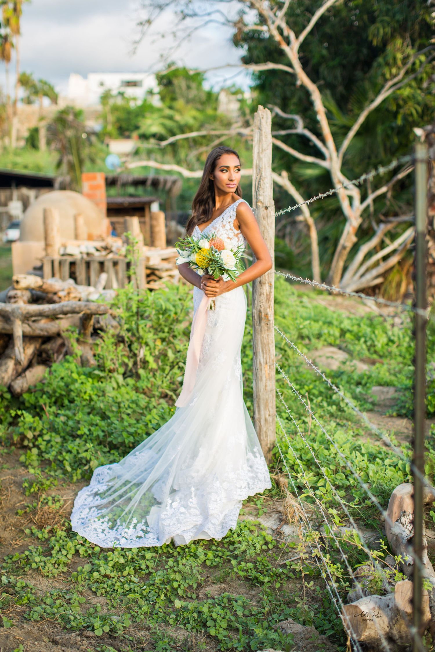 Bride in fitted lace wedding dress with train holding bouquet standing by fence post in garden.