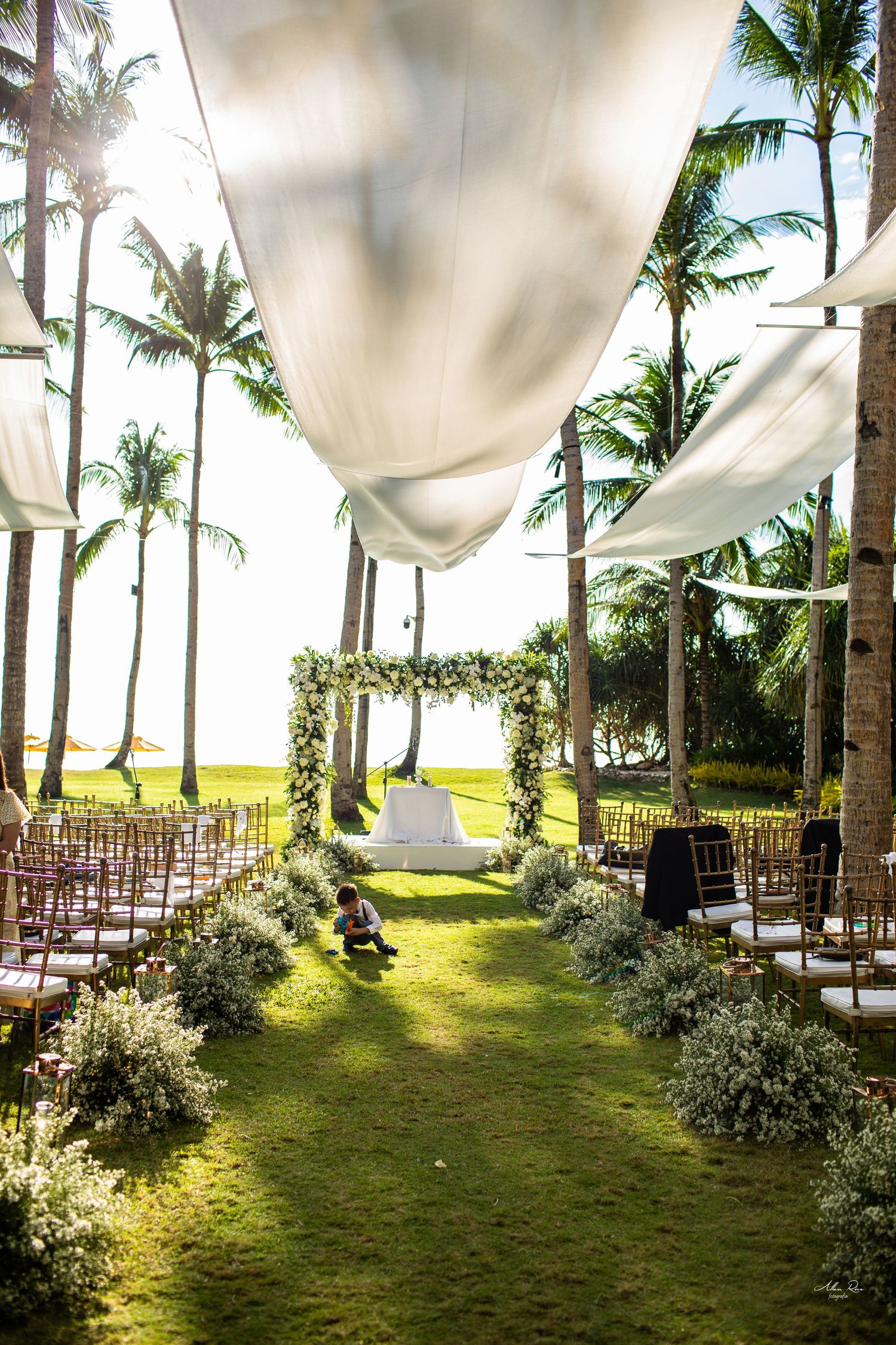 Elegant outdoor wedding ceremony setup with floral arch, palm trees, white draping, and aisle lined with flowers.