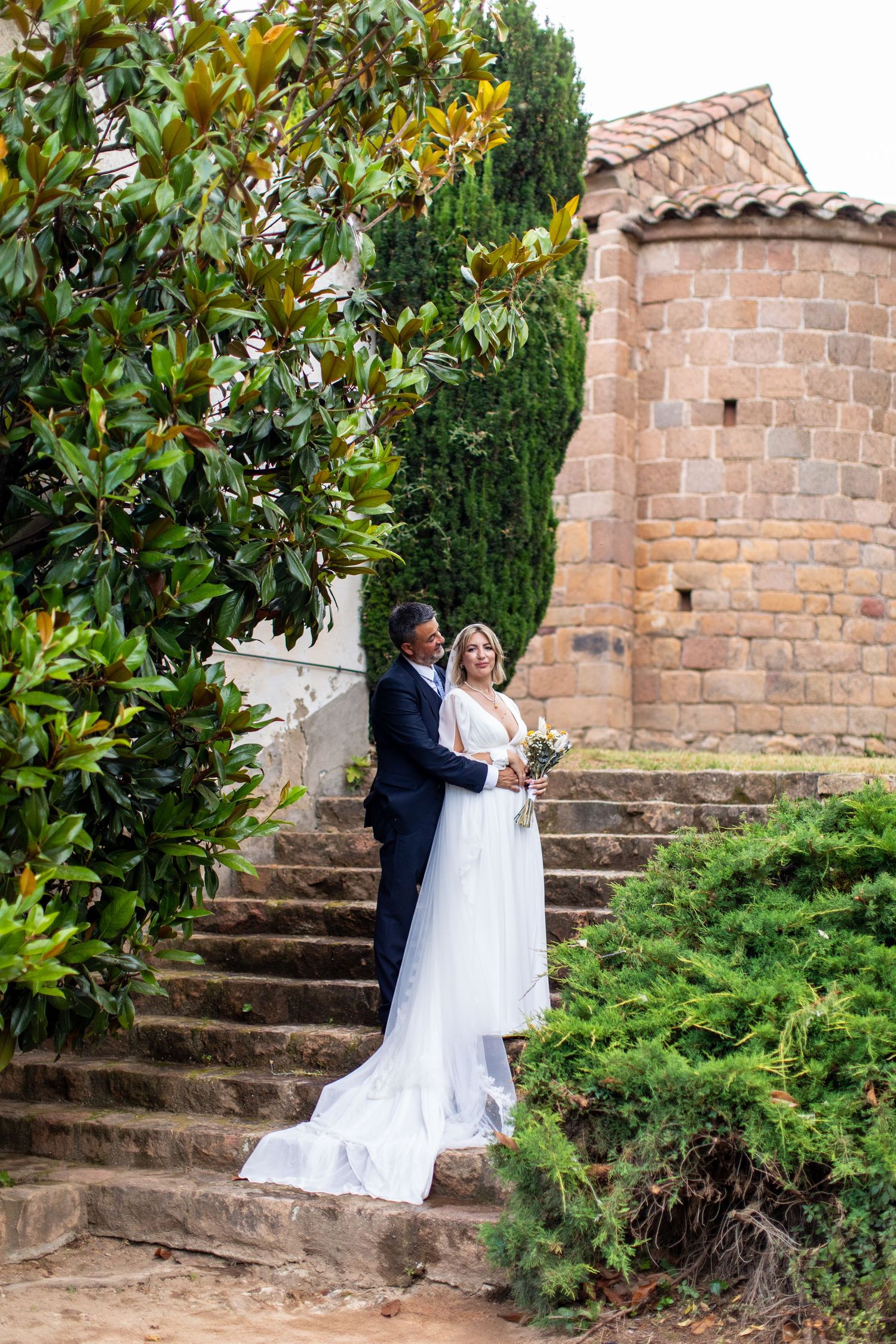 Bride and groom embrace on stone steps beside ancient church surrounded by greenery.