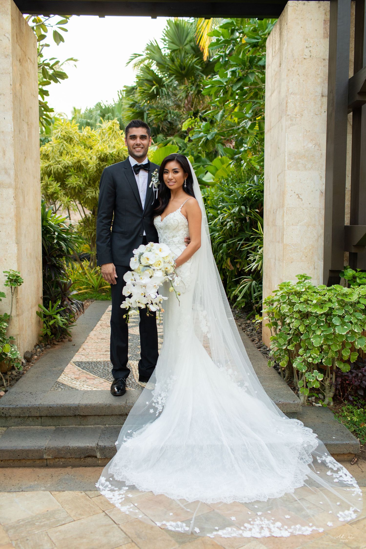 Bride in a lace gown with cathedral veil and groom in a tuxedo pose in a lush tropical garden setting.
