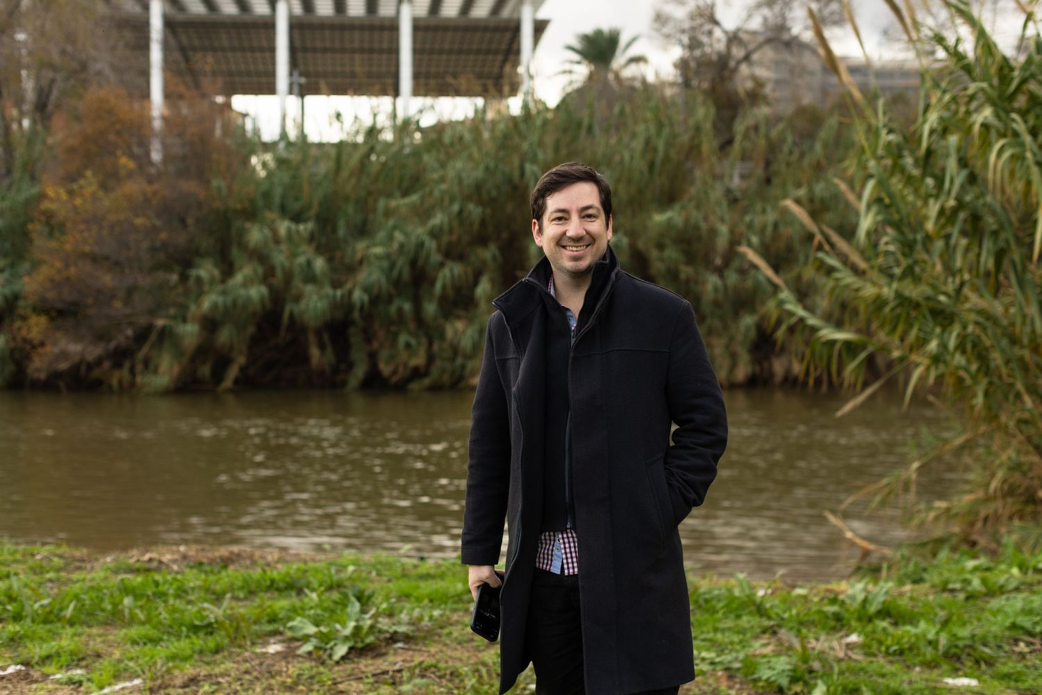 Smiling man in black coat stands outdoors near a river with lush green vegetation behind him.