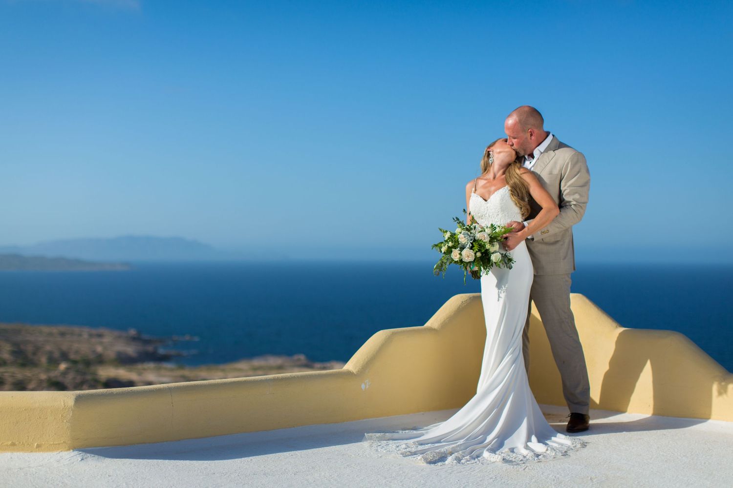 Couple sharing a kiss in wedding attire on white terrace overlooking the blue Santorini sea.