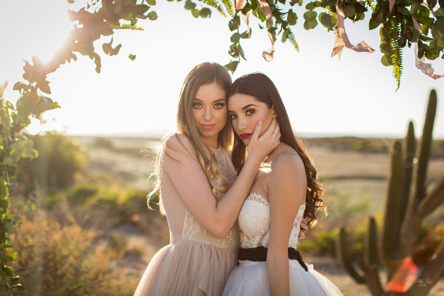 Two brides embracing closely under a leafy arch with cacti and desert dunes behind them.