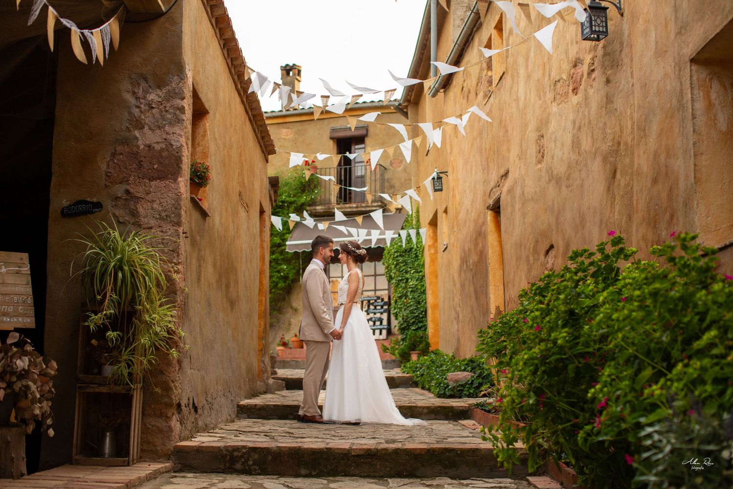 Newlyweds embrace in charming rustic alleyway decorated with bunting flags and lush greenery.