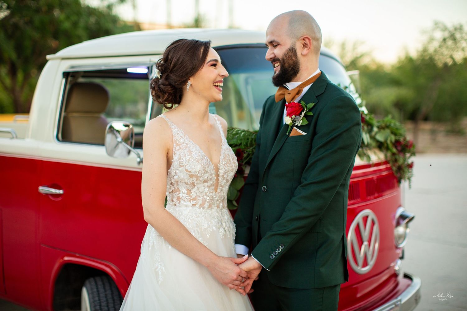 Smiling bride and bearded groom holding hands in front of classic red Volkswagen wedding bus.