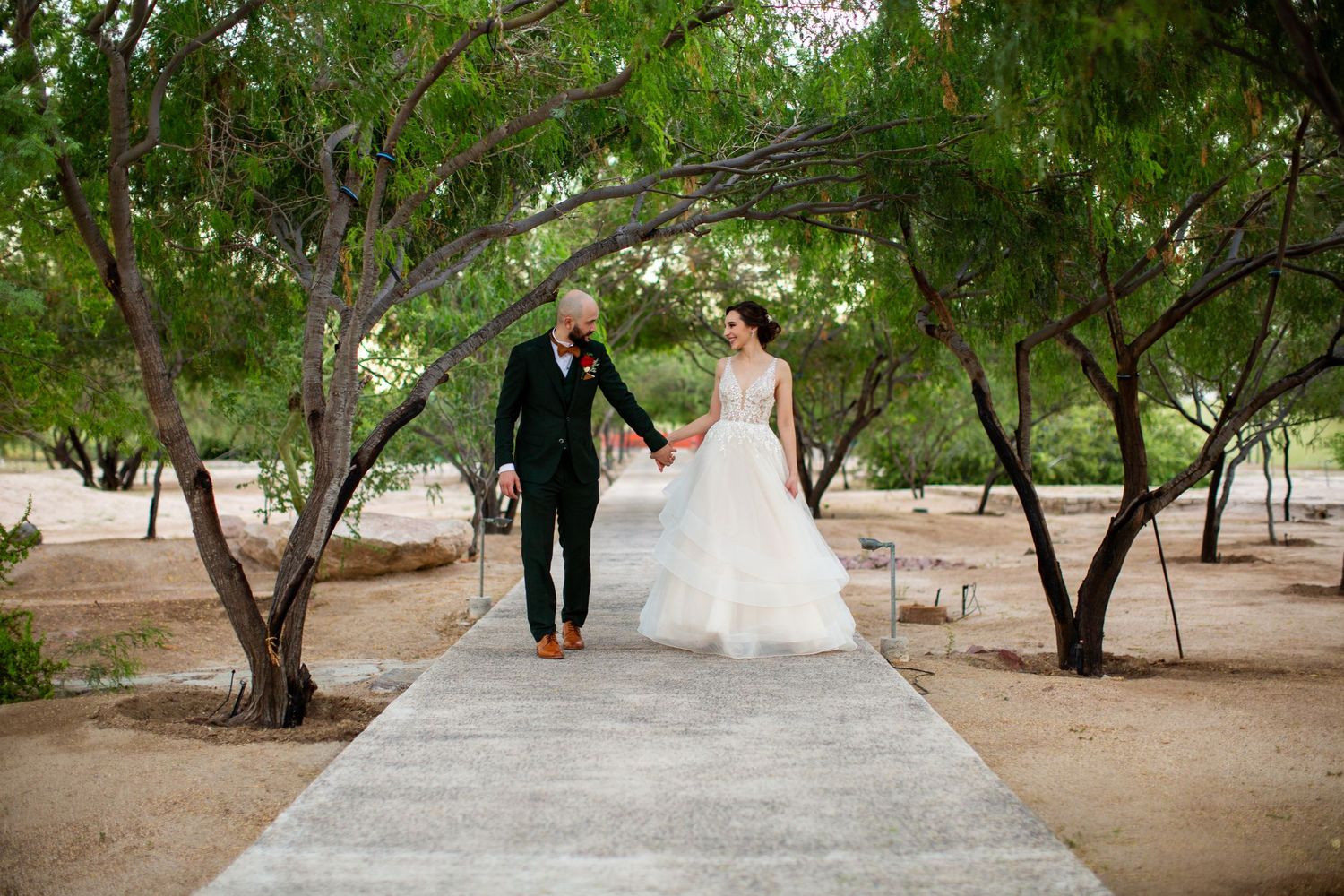 Bride and groom walking hand in hand along tree-lined pathway at outdoor wedding venue.