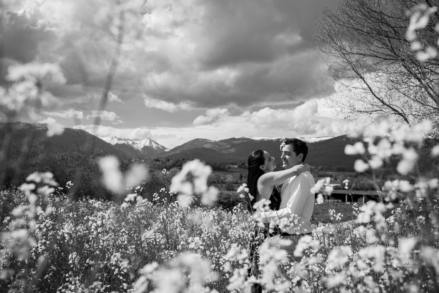 Couple embracing in field of white wildflowers with mountain backdrop in black and white photo.