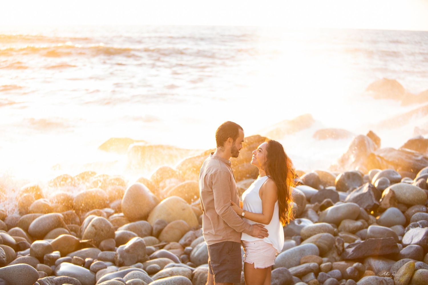 Couple kissing on a rocky beach at sunset with golden waves crashing behind them.