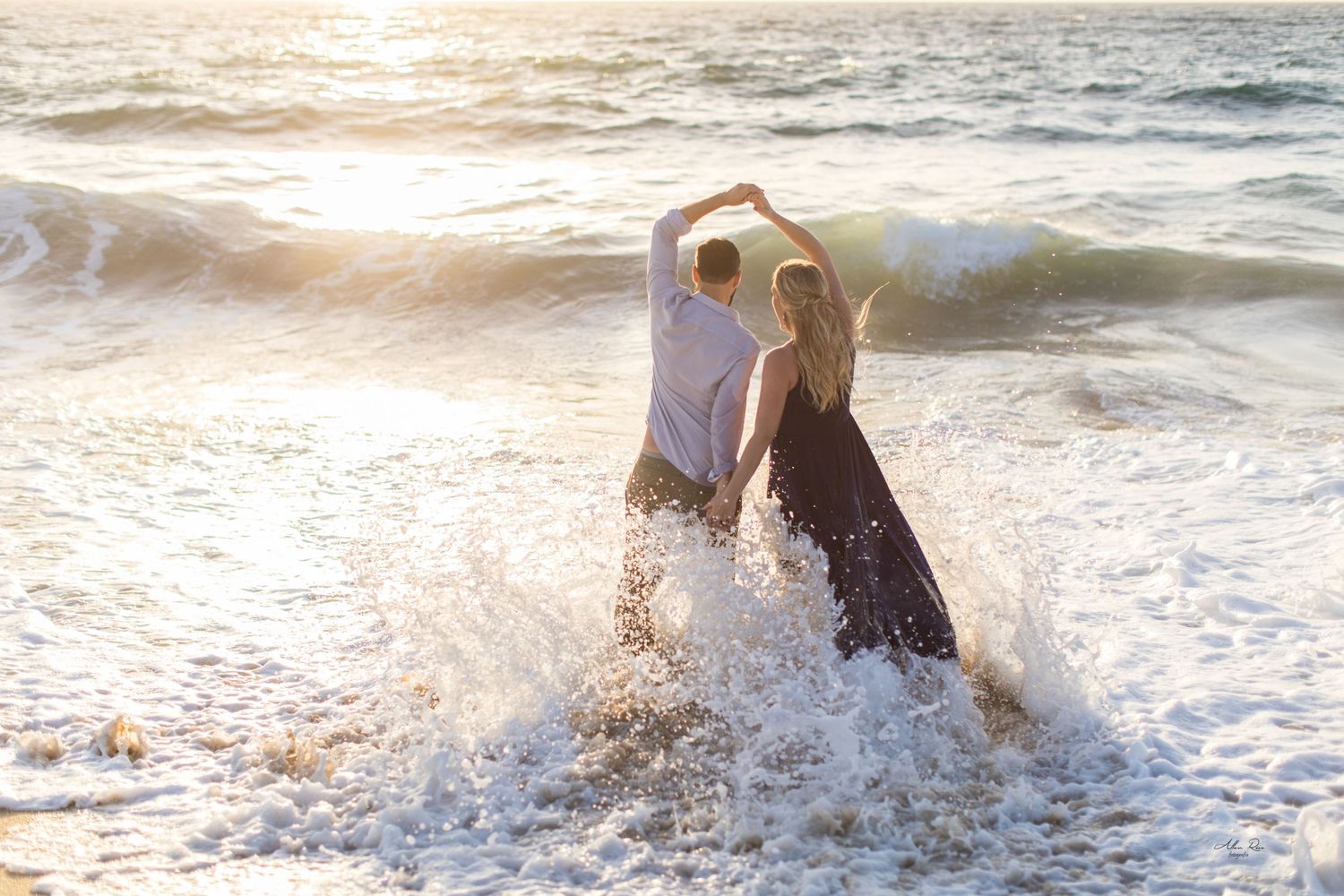 Couple dancing joyfully in ocean waves at sunset, splashing water in a flowing blue dress.