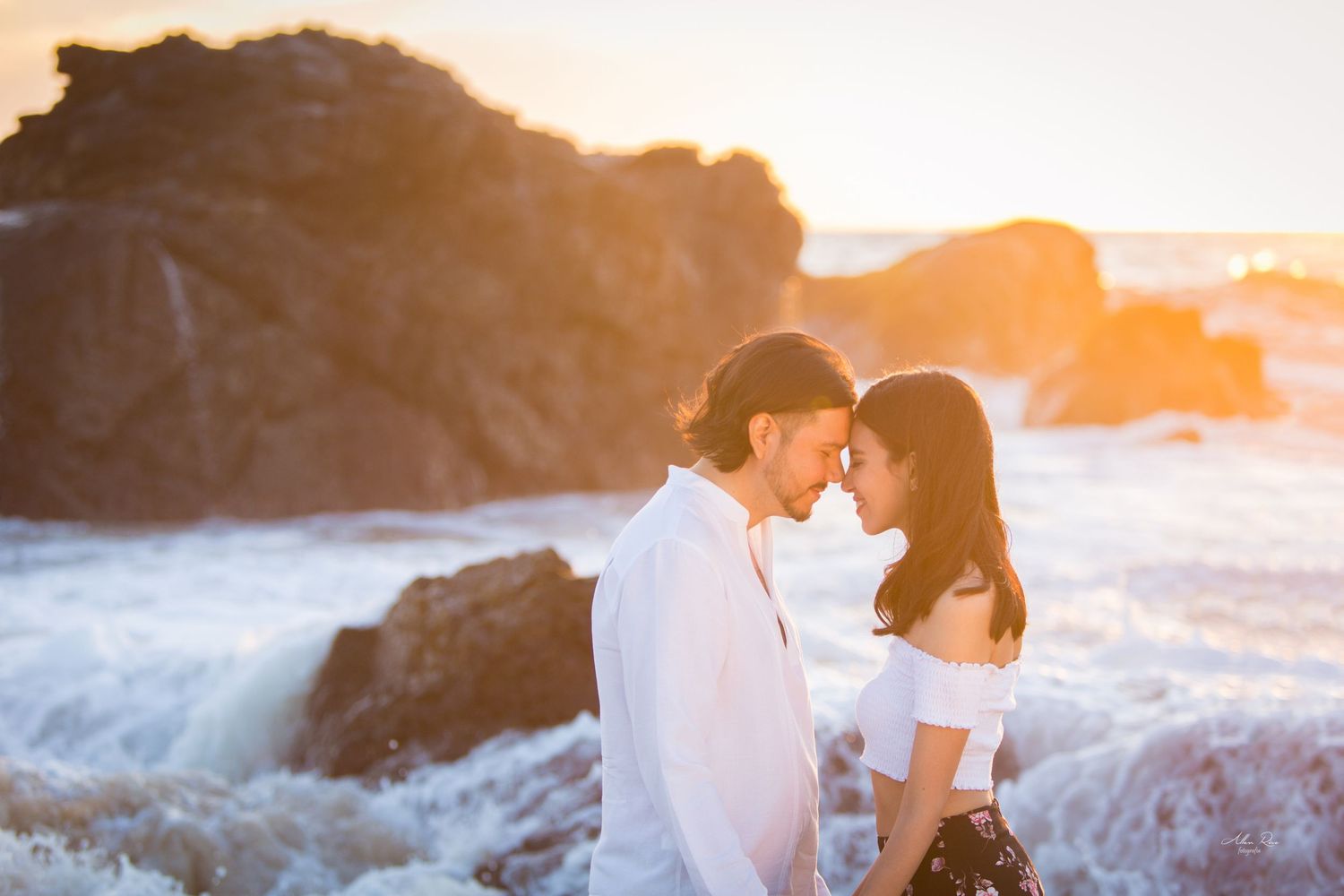 Romantic couple touching foreheads near crashing ocean waves at golden sunset.