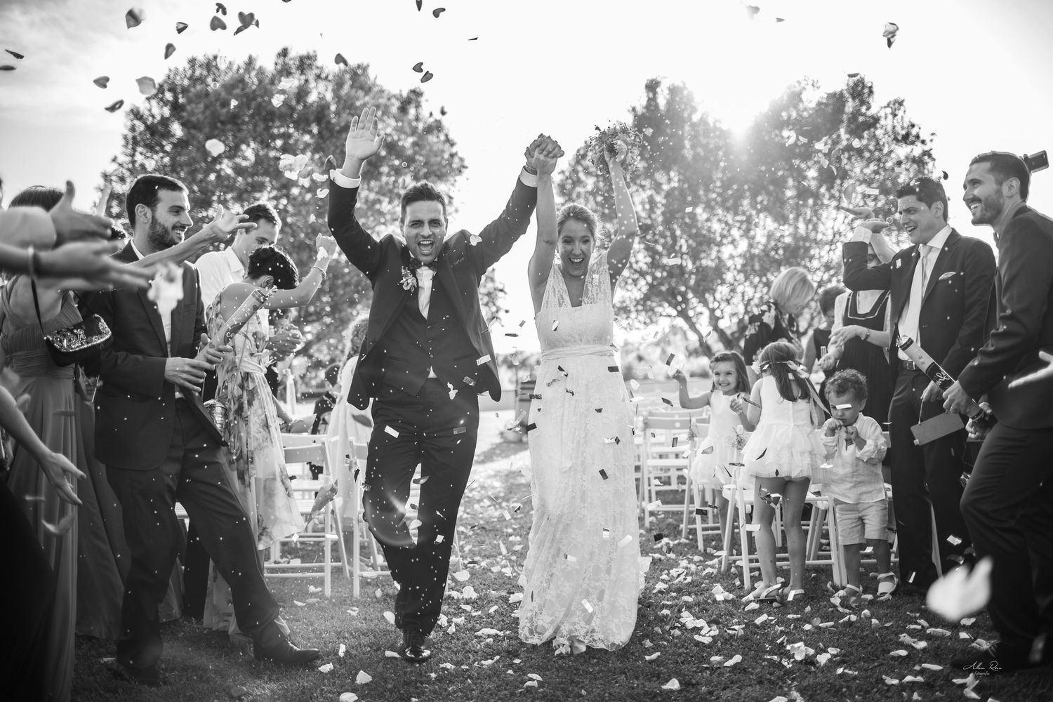 Black and white photo of newlyweds celebrating recessional with confetti and cheering guests outdoors.