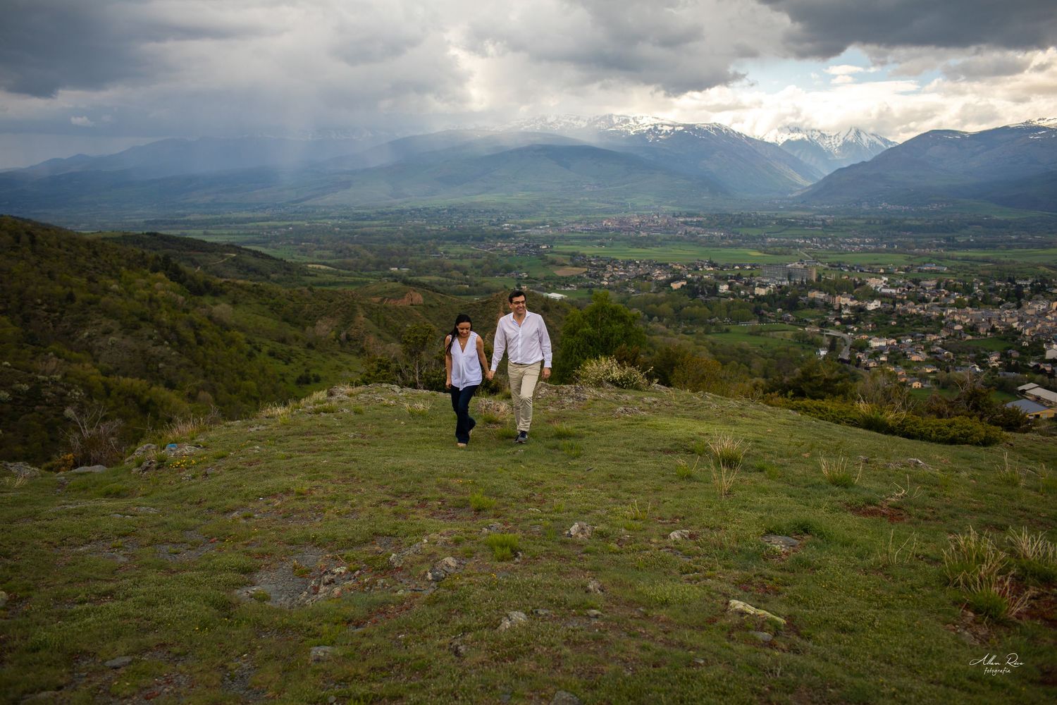 Couple walking hand in hand on a hilltop meadow with a stunning panoramic valley and mountain view.