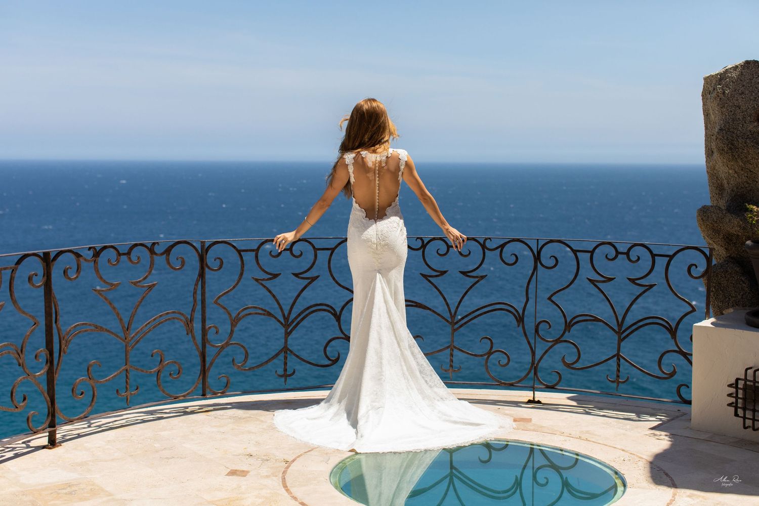 Bride in backless white mermaid gown stands at ornate iron balcony railing overlooking stunning blue ocean view.