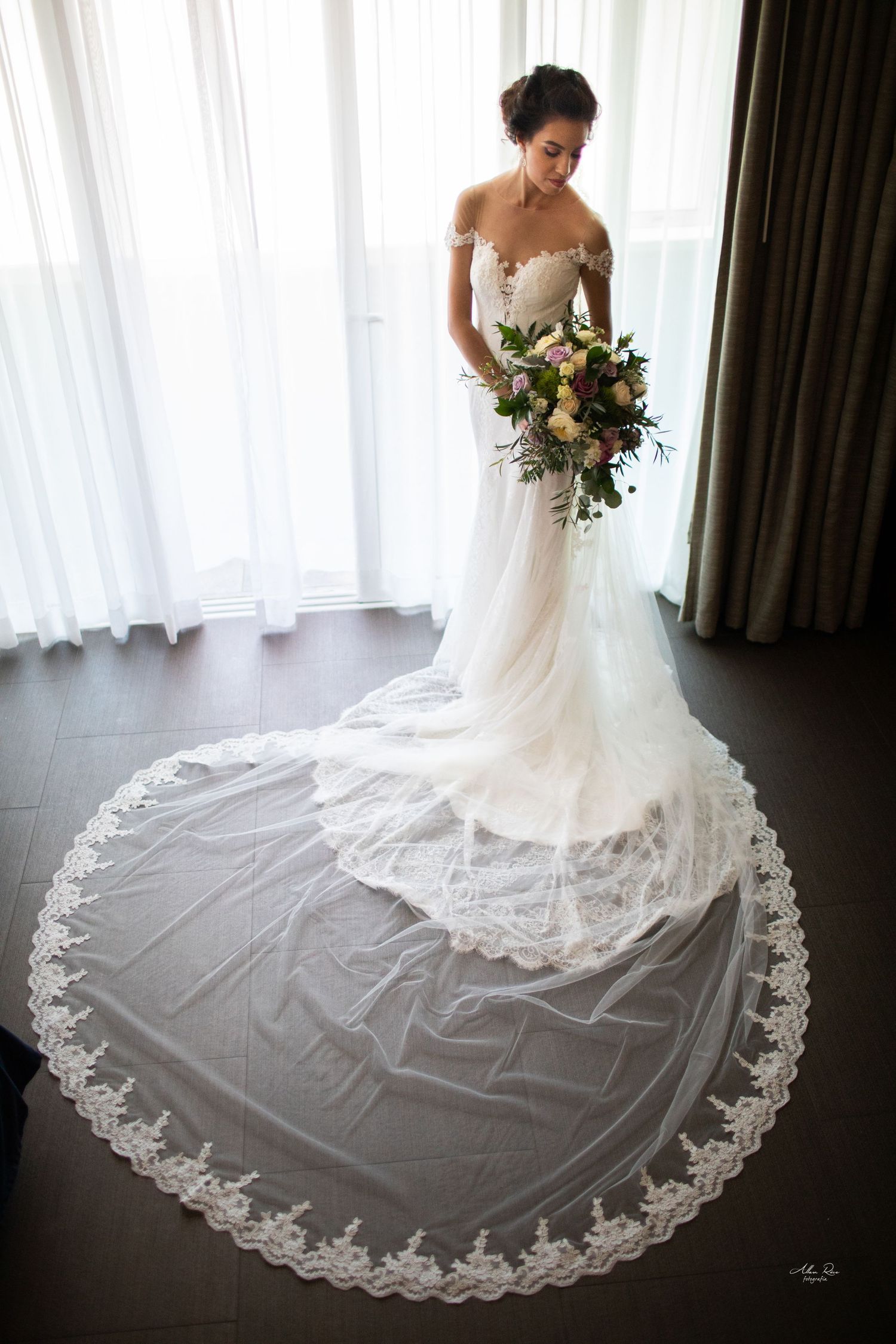Bride in off-shoulder lace gown with cathedral veil spread on floor holding a colorful bridal bouquet.