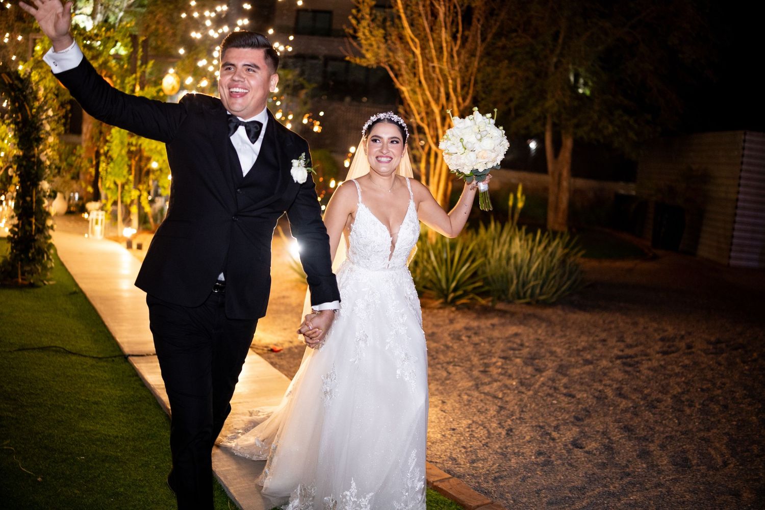 Happy bride and groom celebrating at night under string lights, bride holding white bouquet.