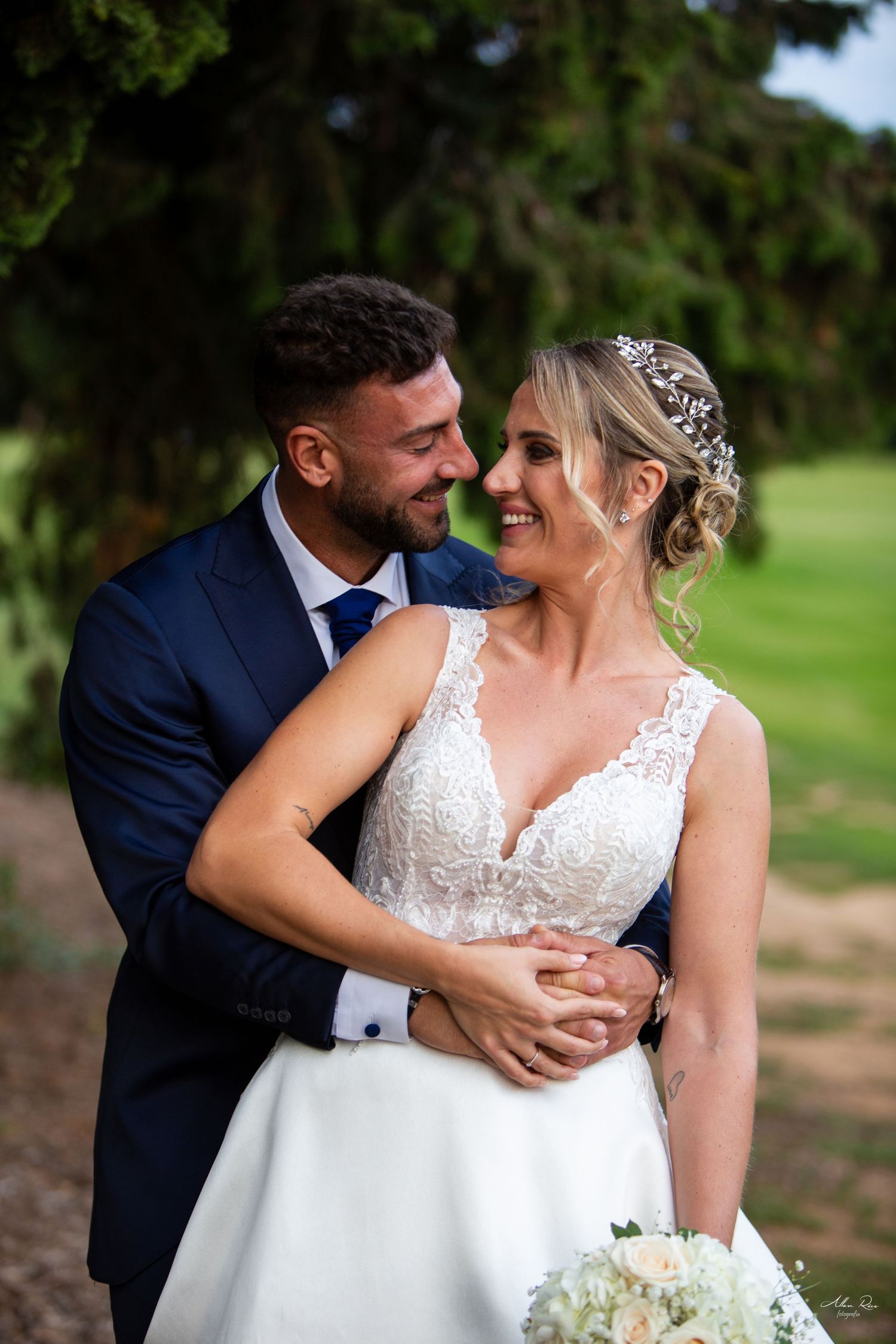 Groom embraces smiling bride in lace wedding gown as they share a tender romantic moment outdoors.