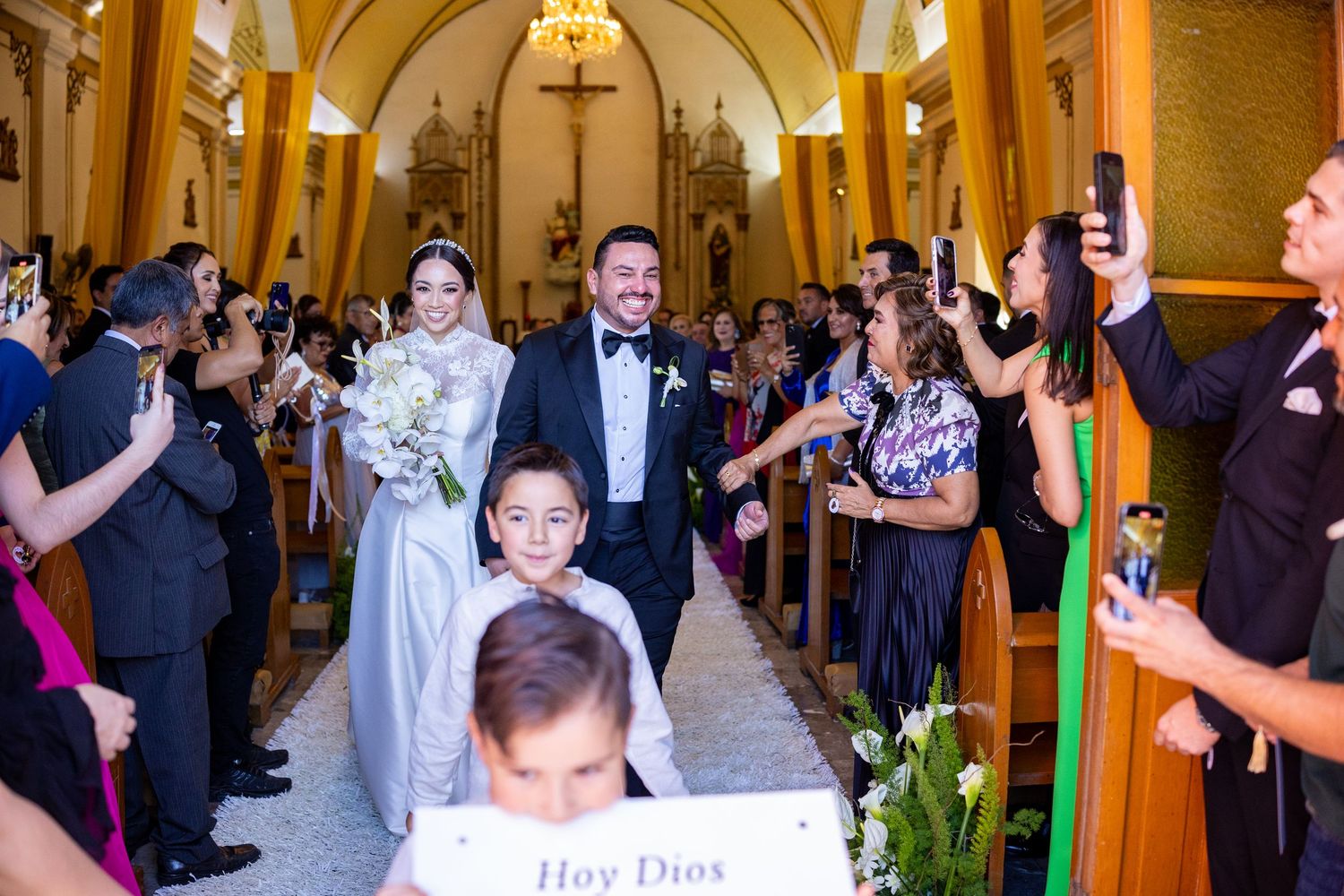 Bride and groom walking down church aisle with flower girl holding sign at wedding.