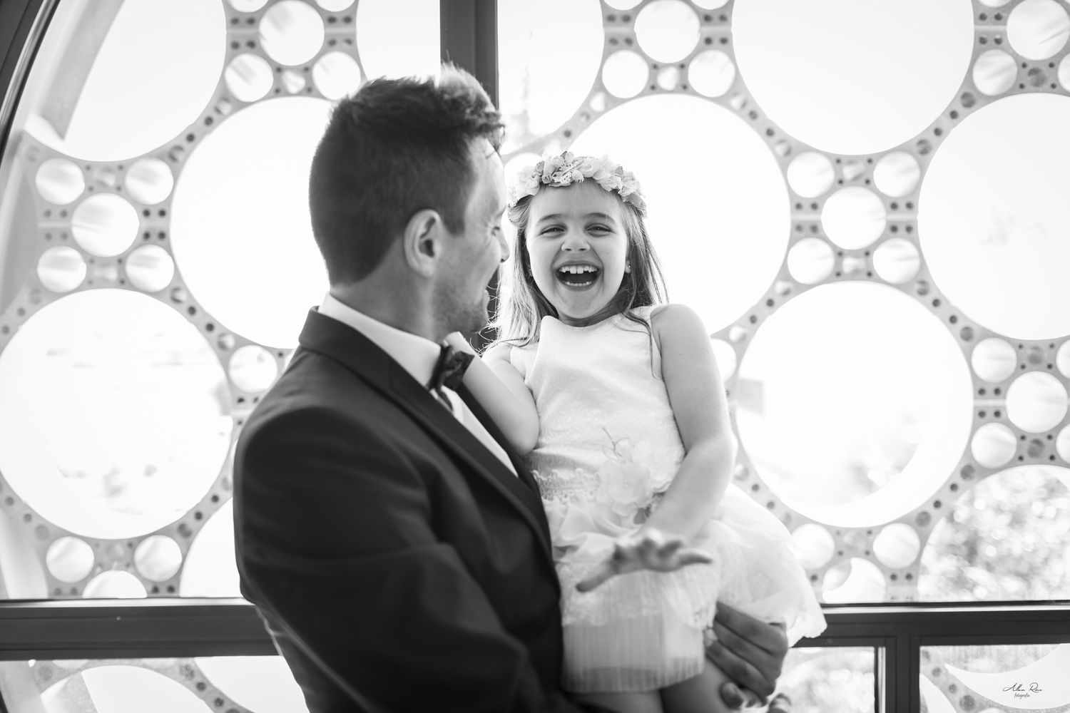 Black and white photo of groom holding laughing flower girl by ornate window.