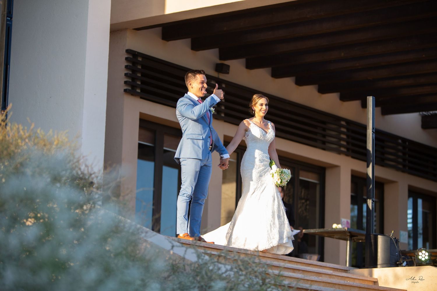Bride and groom stand elegantly on a terrace overlooking a scenic outdoor venue in matching blue and white.