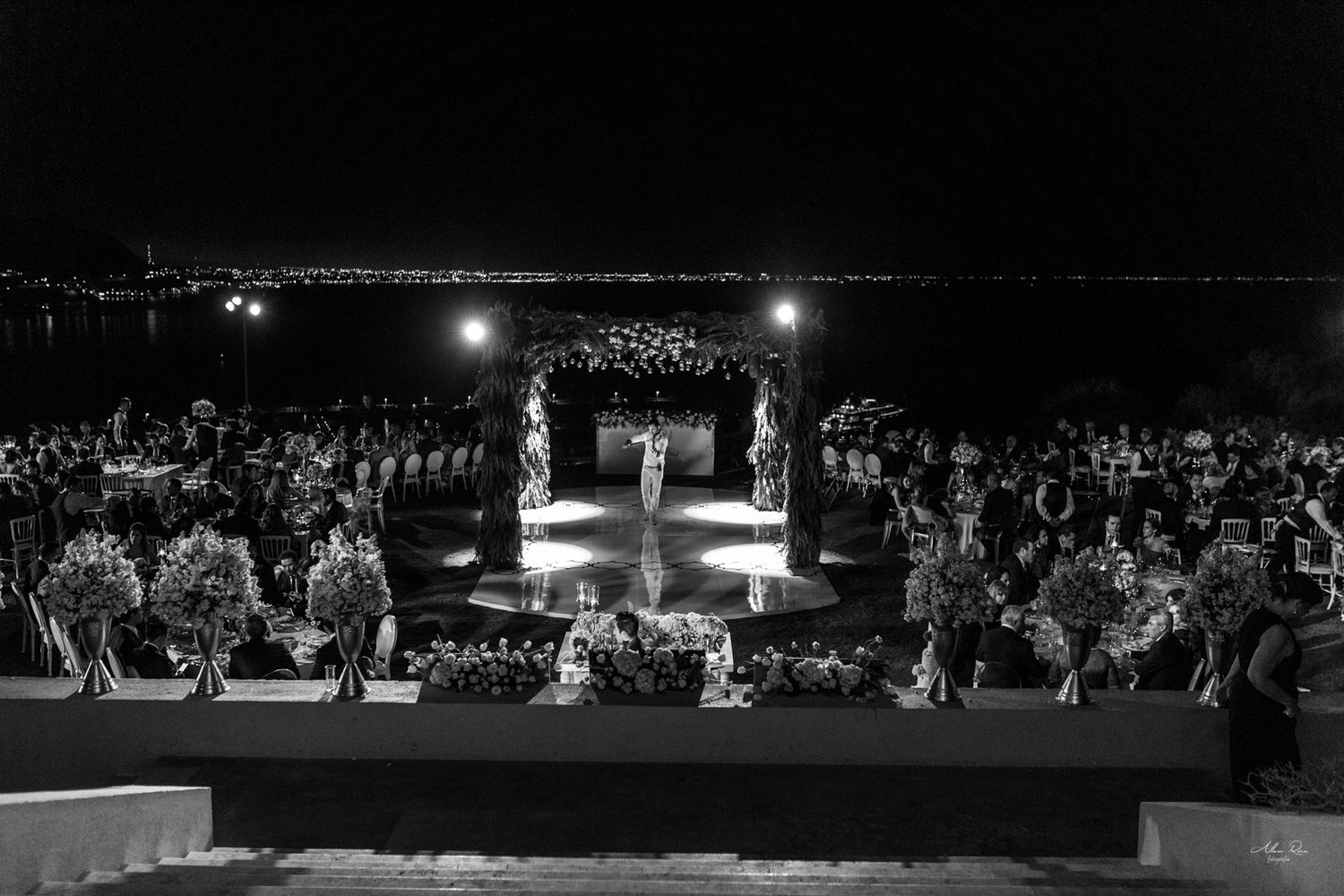 Black and white aerial view of elegant outdoor nighttime wedding ceremony with floral arch by the water.