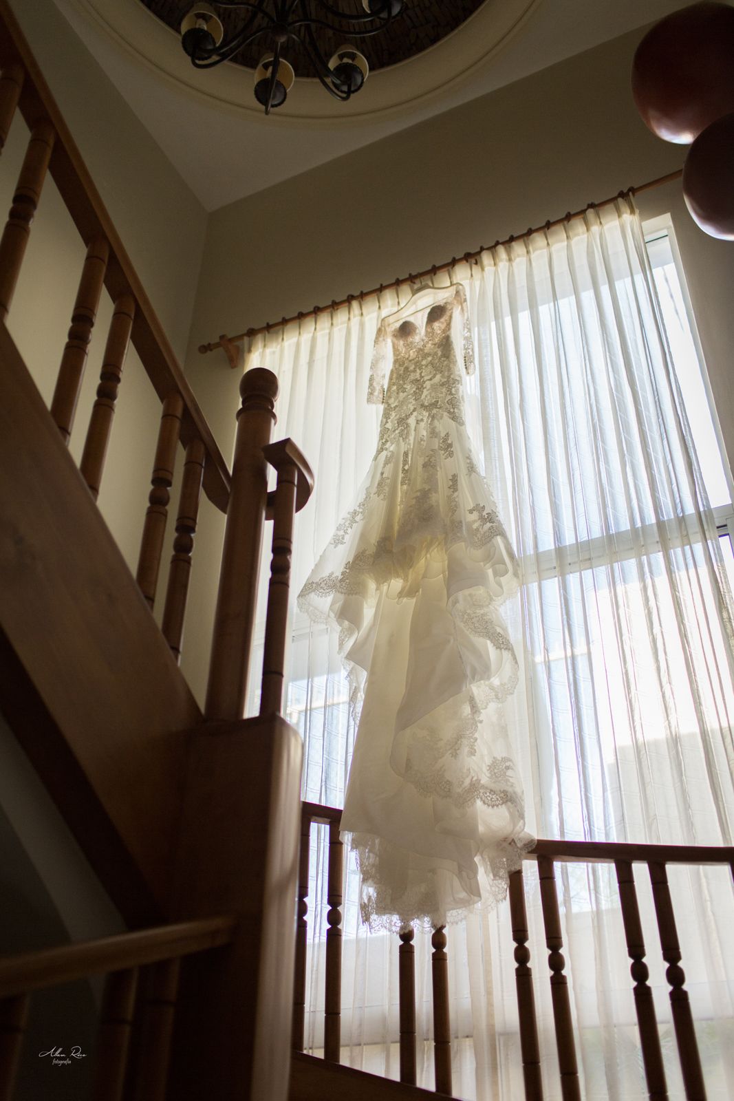 Elegant wedding dress hanging by a staircase window with sheer curtains and natural light.