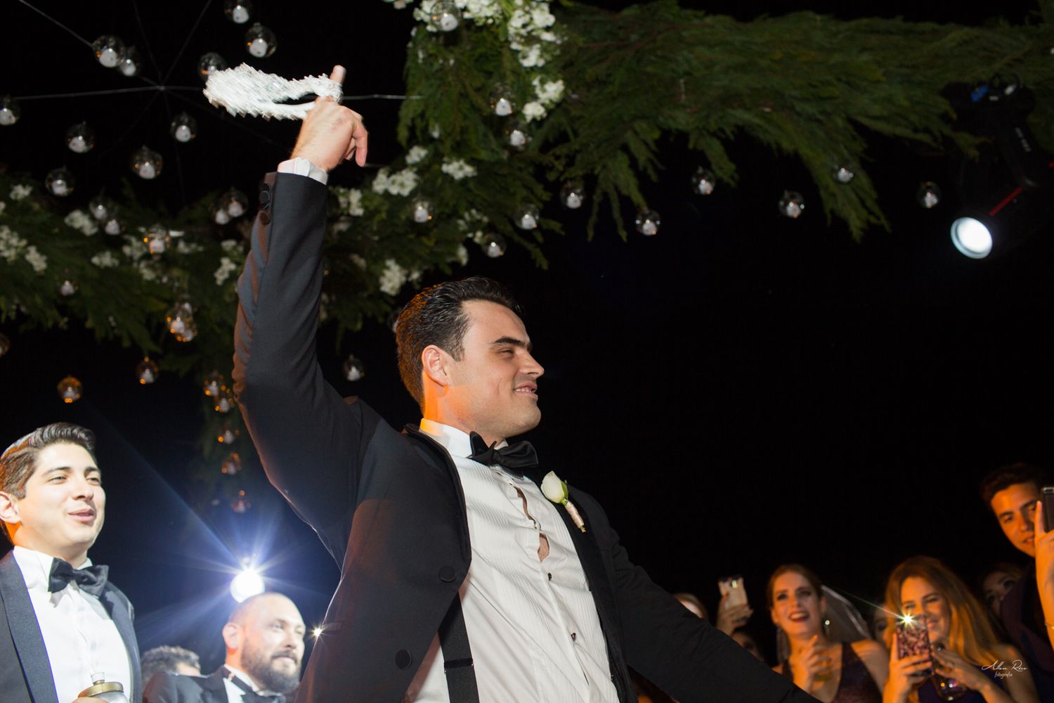 Groom raising bouquet triumphantly at outdoor nighttime wedding celebration.